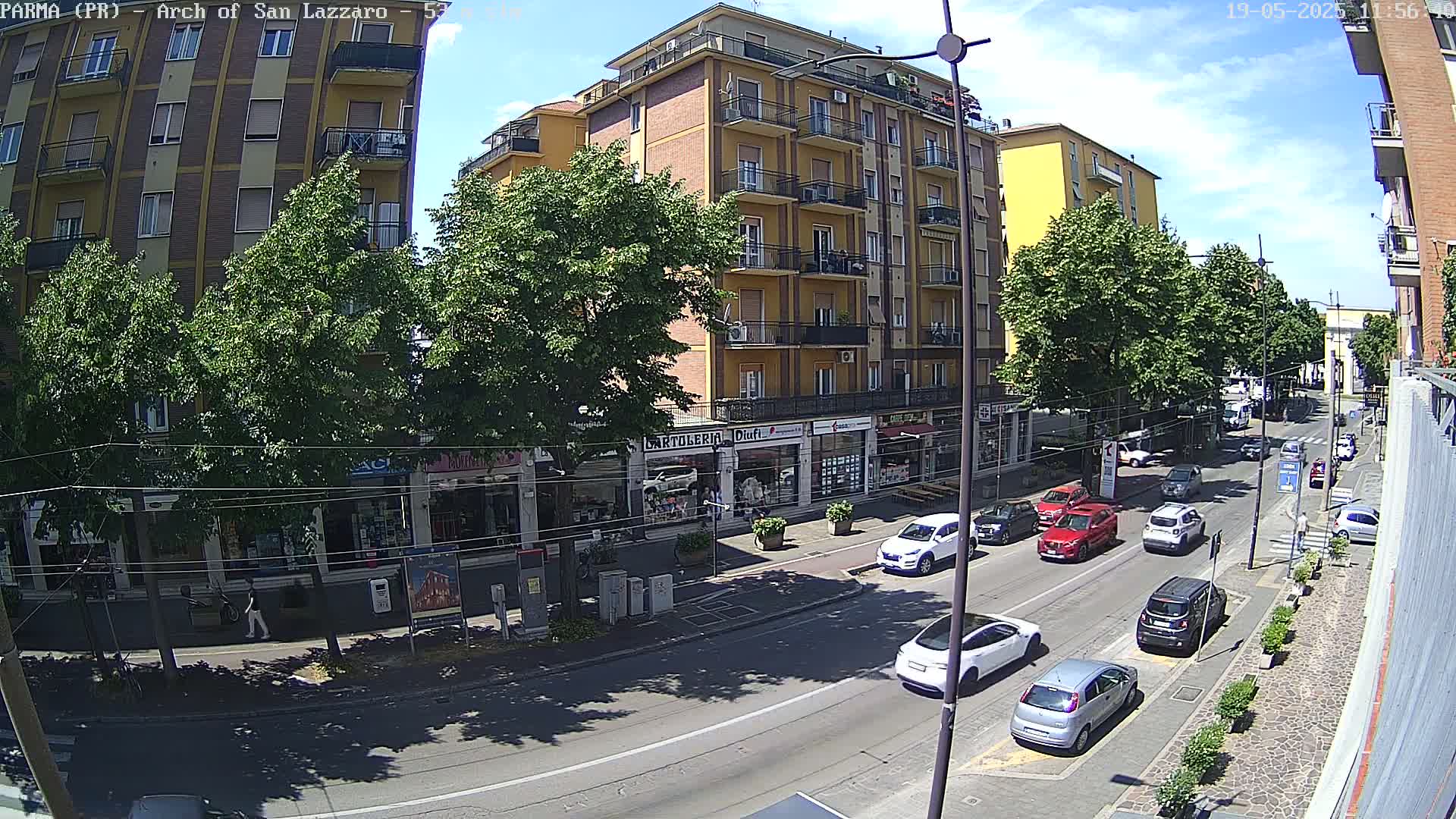A sunny day on a city street shows several cars driving past a multi-story building with shops on the ground floor, lined with trees and pedestrians.