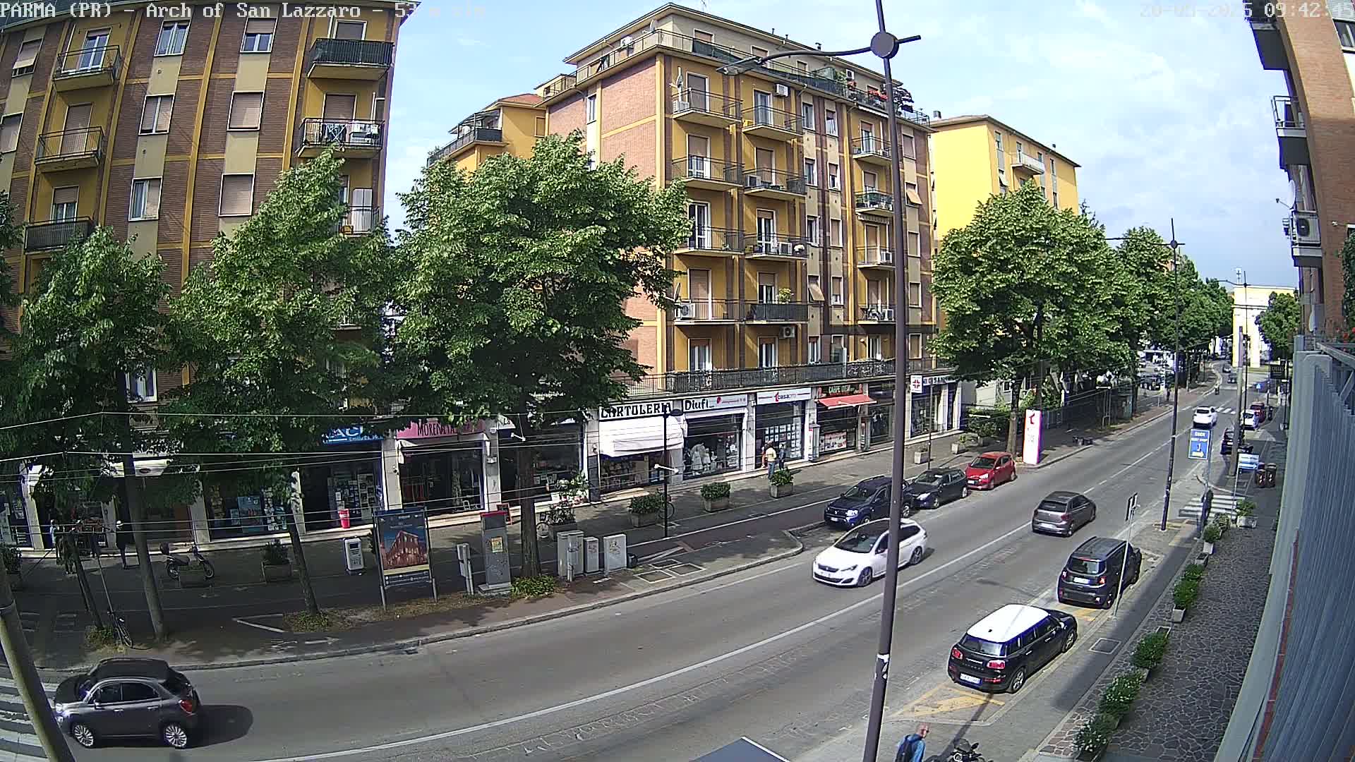 A sunny street scene shows a multi-story yellow building with shops on the ground floor, lined with trees, and several cars parked and driving on a road.