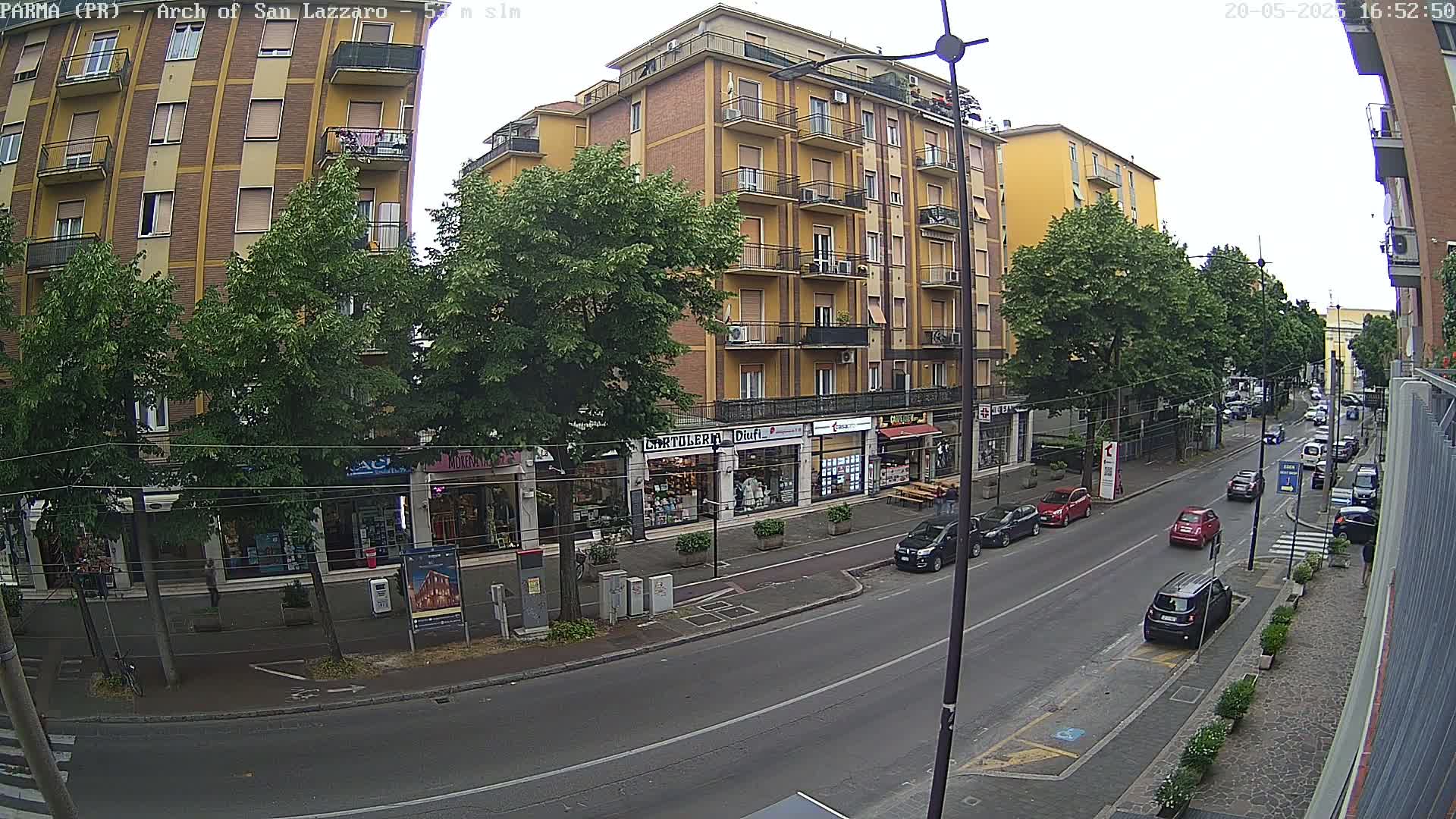 A street scene on an overcast day shows a multi-story yellow building with shops on the ground floor, lined with trees, and several cars parked and driving along the street.
