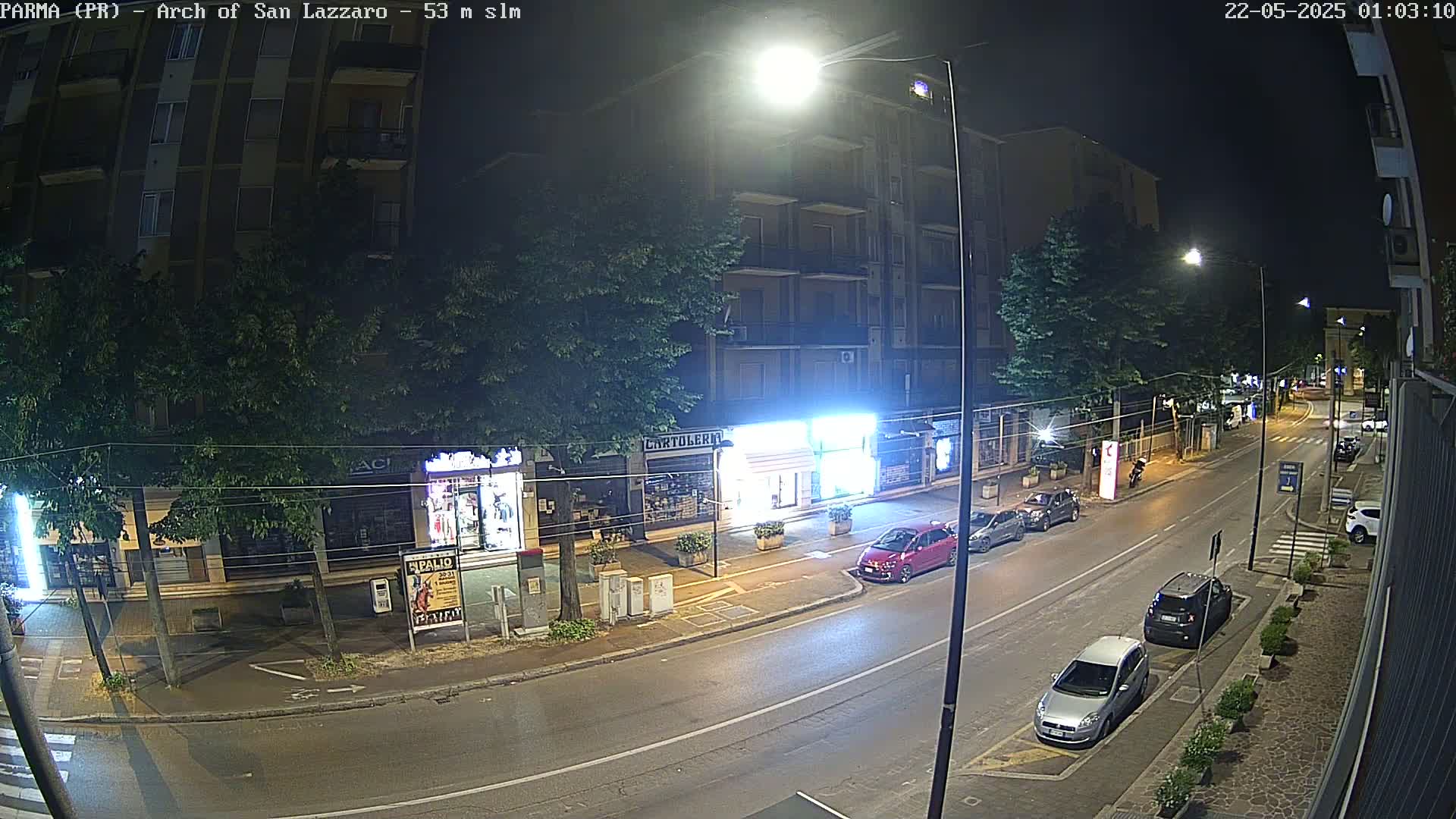 A nighttime street scene shows a mostly empty road with several parked cars, illuminated by streetlights, in front of multi-story buildings and businesses.