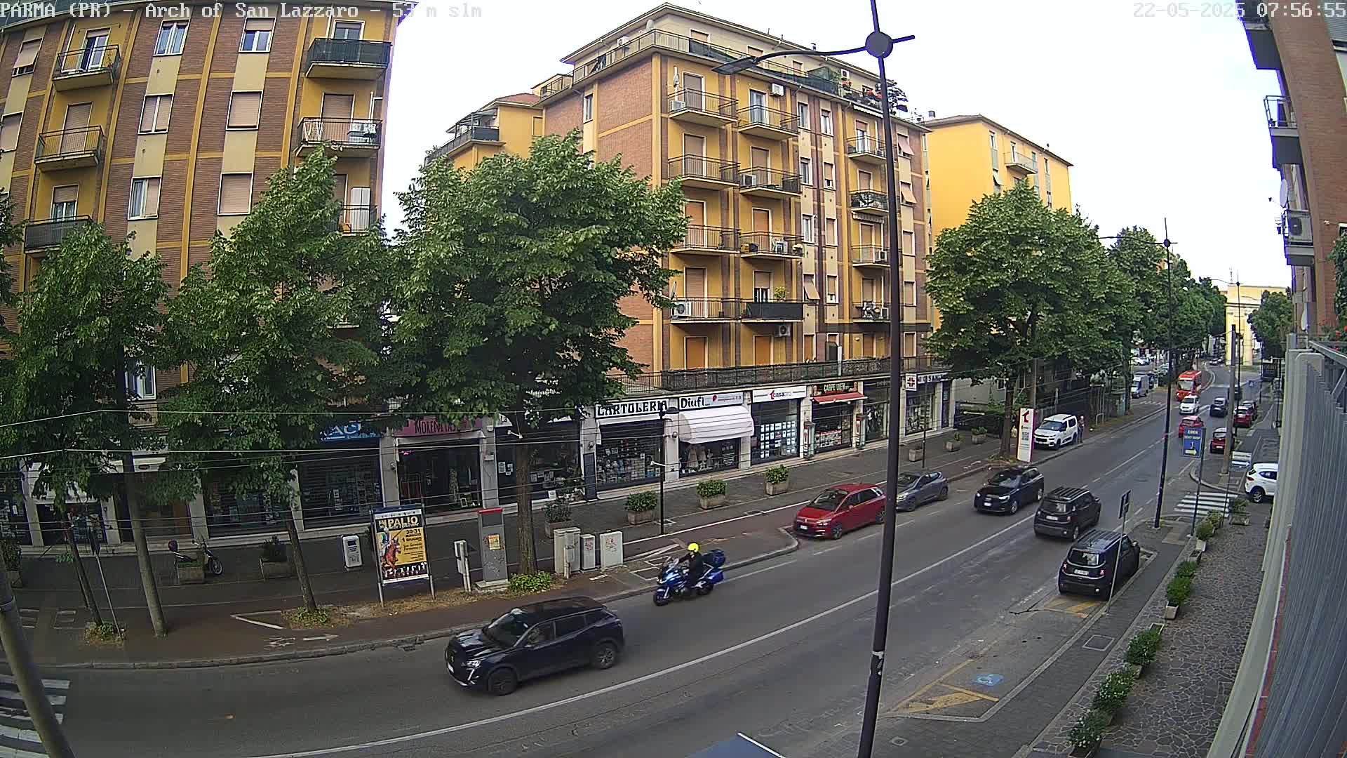 A street scene on an overcast day shows a multi-story yellow building with shops on the ground floor, lined with trees, and several cars and a motorcycle on the road in front.