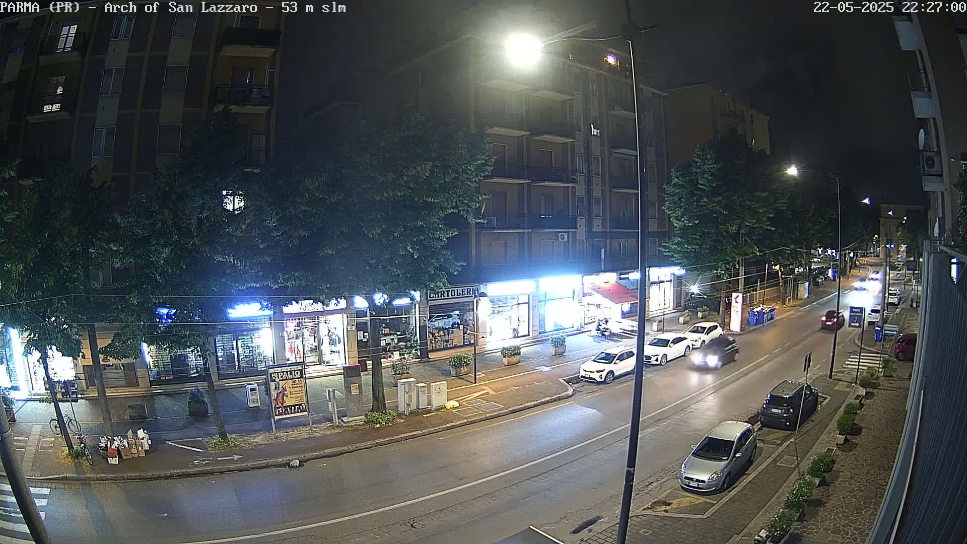 A nighttime street scene shows several parked and moving cars on a road lined with shops and buildings, under streetlights.