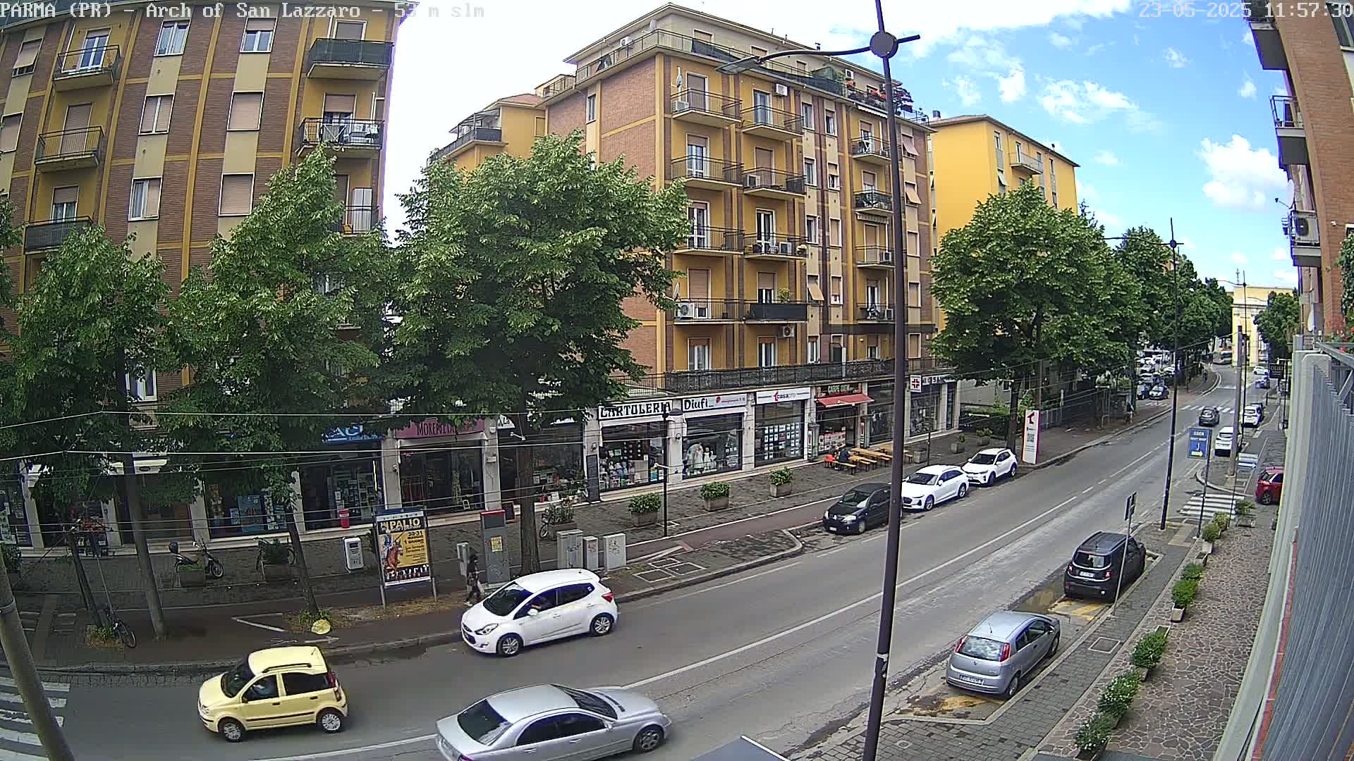 A street scene on a partly sunny day shows parked cars and several multi-story buildings, one large yellow building prominent in the view.