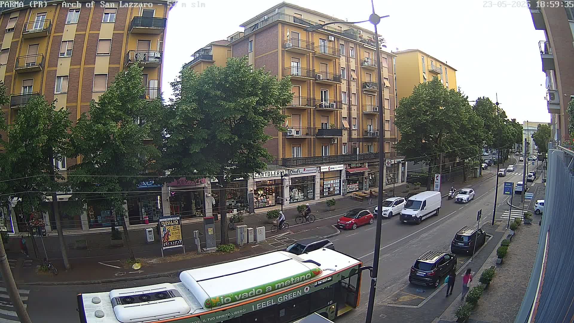A city street scene under an overcast sky shows several cars, a bus, and pedestrians near multiple-story buildings with shops on the ground floor and trees lining the street.