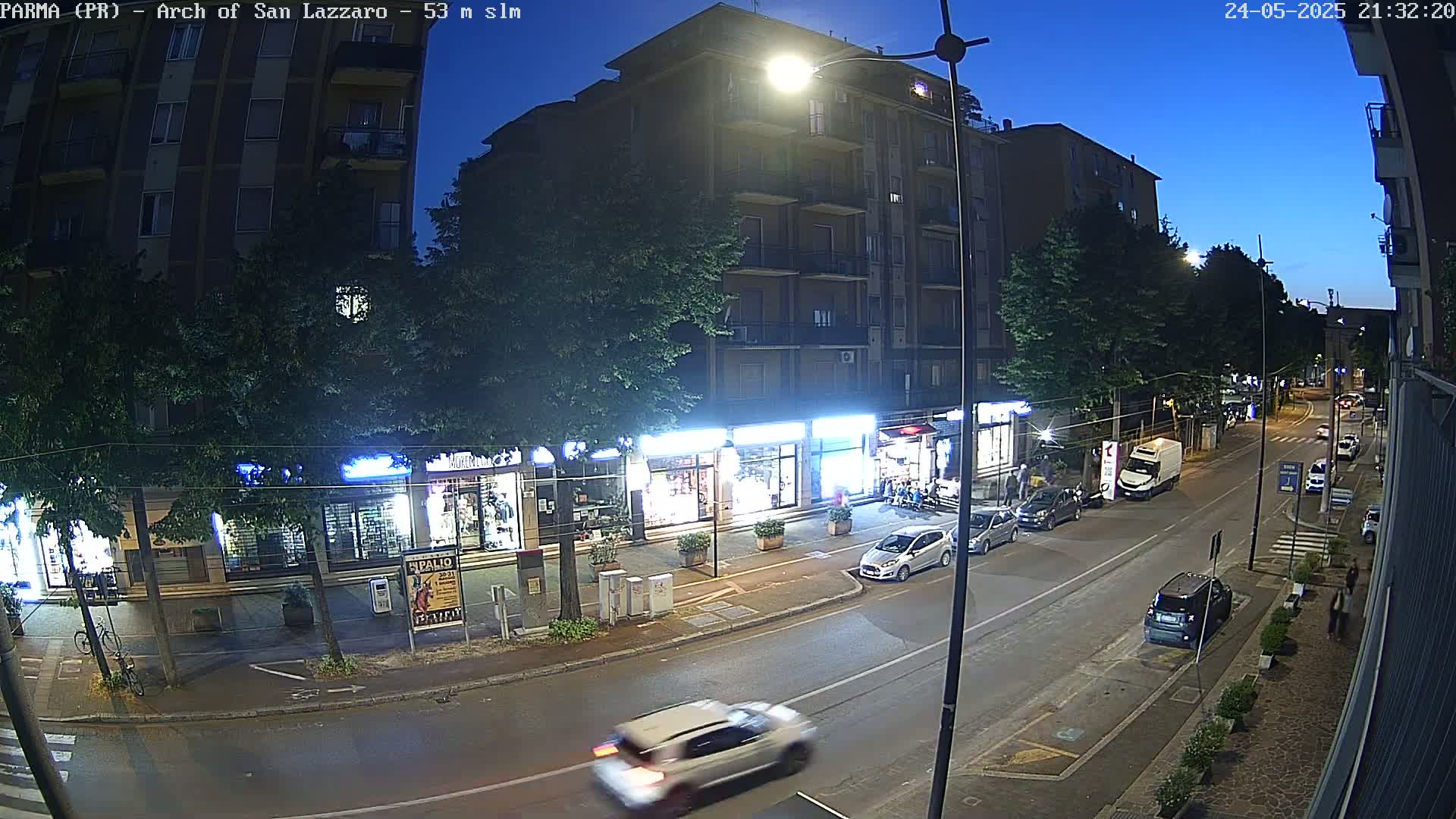 A nighttime street scene shows a multi-story building and shops under a twilight sky, with several cars parked and driving along the road.