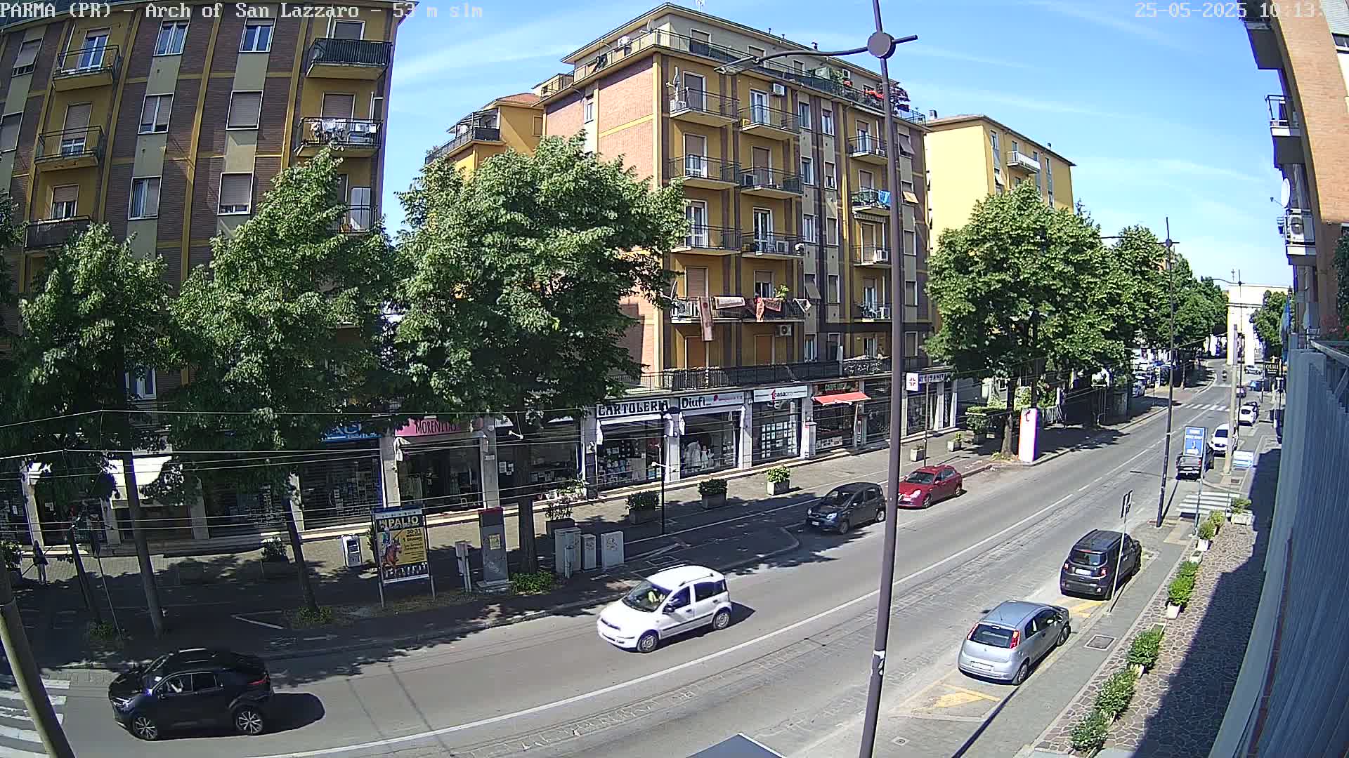 A sunny city street scene shows a multi-story yellow building with shops on the ground floor, several cars parked and driving on the road, and green trees lining the sidewalks.