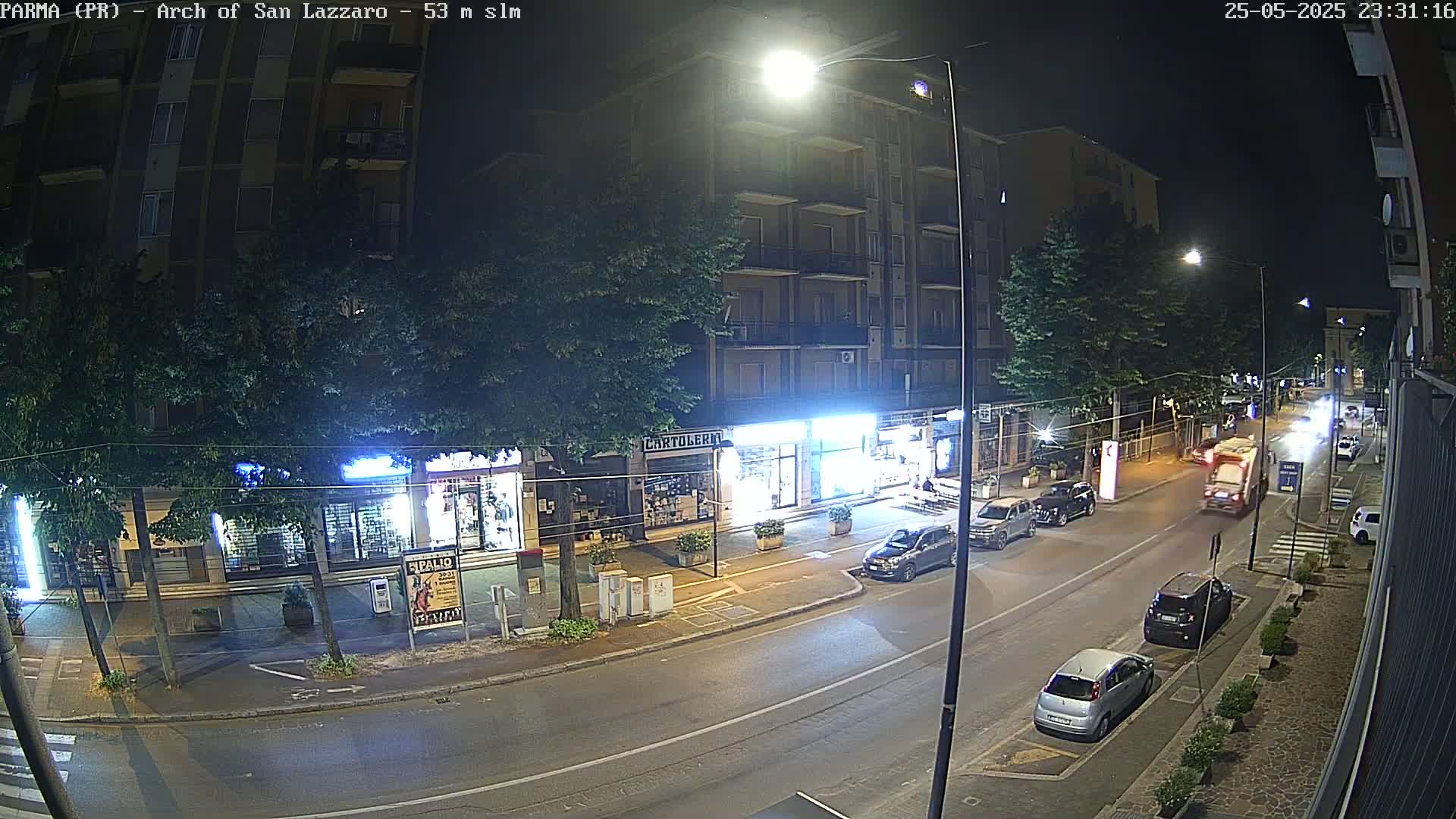 A nighttime street scene shows several cars parked and driving along a road lined with shops and apartment buildings under bright streetlights.