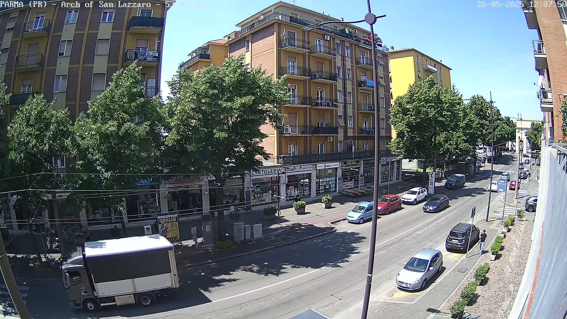 A sunny city street scene shows a multi-story building with shops on the ground floor, several cars parked along the side of the road, and a truck driving down the street.