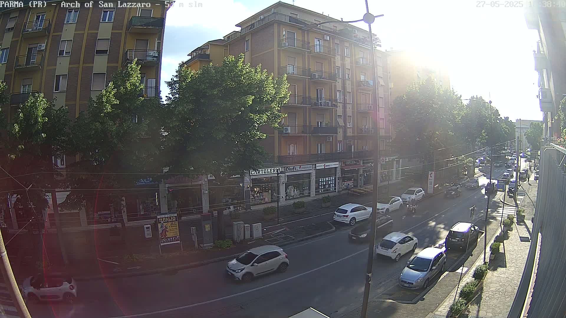 A sunny street scene shows a multi-story building with shops on the ground floor, several cars parked and driving along the street, and lush green trees lining the sidewalk.