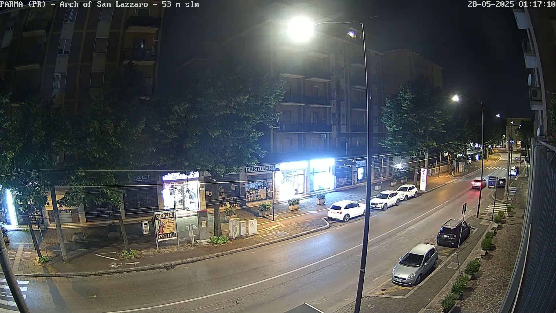 A nighttime street scene shows several parked cars and a few driving cars along a road lined with buildings and trees under bright streetlights.