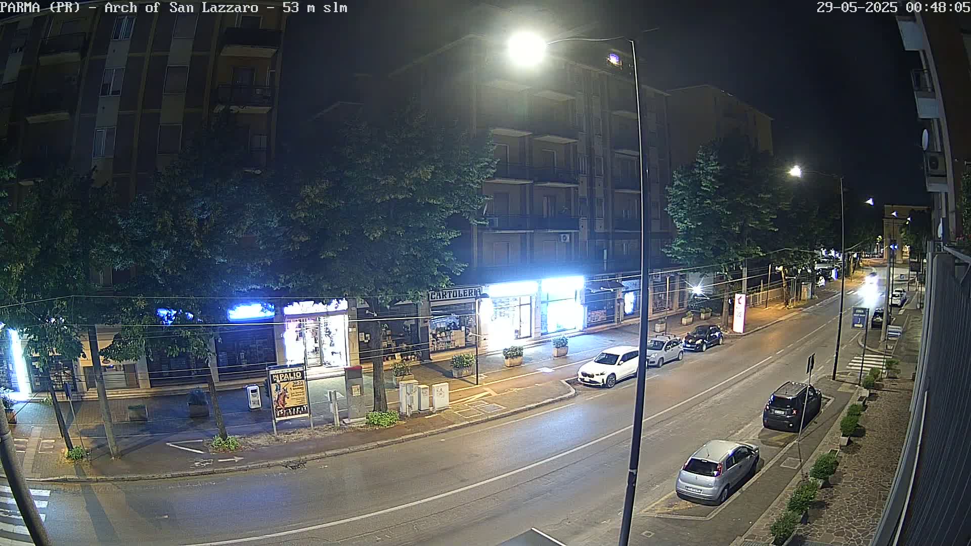 A nighttime street scene shows a multi-story building with street-level shops, several parked cars, and a few cars driving down the road.