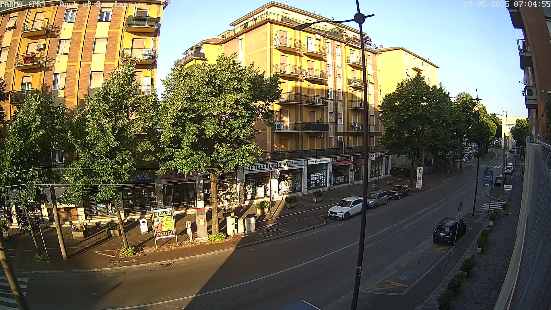 A sunny street scene shows a multi-story yellow building with shops on the ground floor, lined with leafy green trees, and several cars parked along the road.