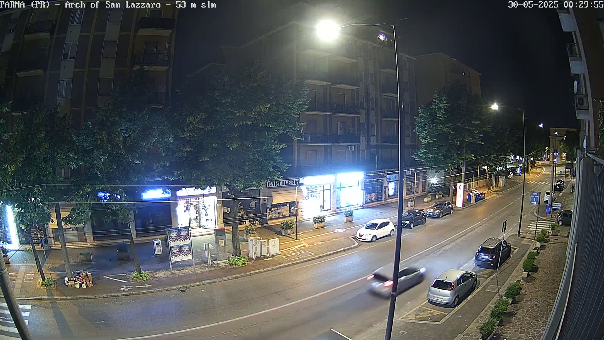 A nighttime street scene shows a multi-story building and several parked and moving cars under bright streetlights.