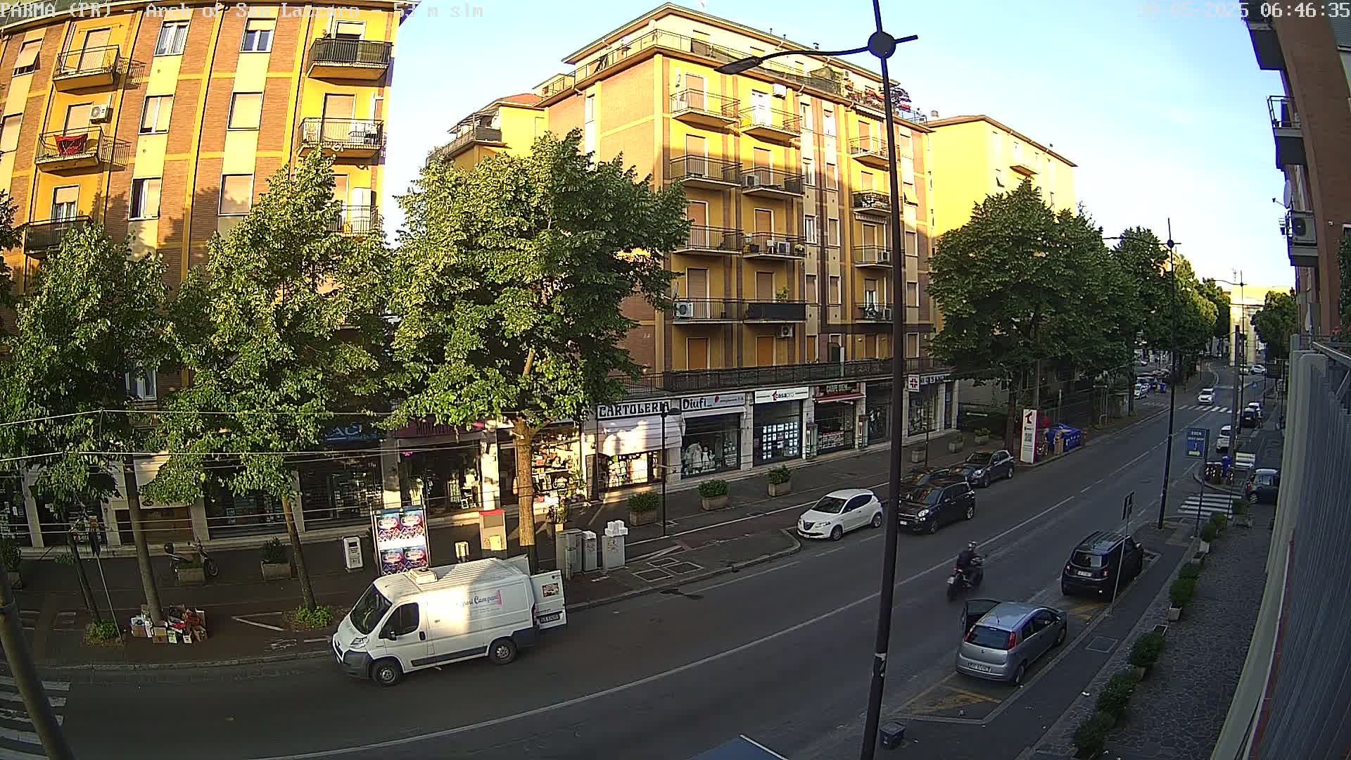A sunny street scene shows a row of yellow buildings with shops on the ground floor and parked cars lining the sides of a road, with a few cars and motorcycles driving along it.