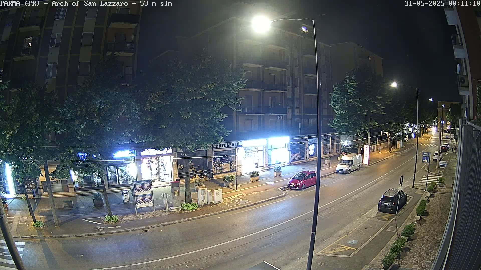 A nighttime street scene shows a mostly empty road lined with shops and buildings under artificial light.