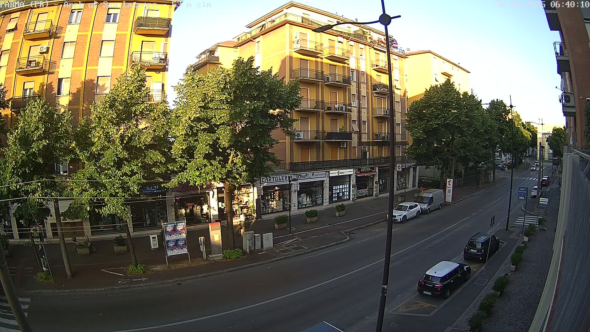 A sunny street scene shows a row of multi-story buildings with shops on the ground floor, lined with green trees, and several cars parked along the side of the road.