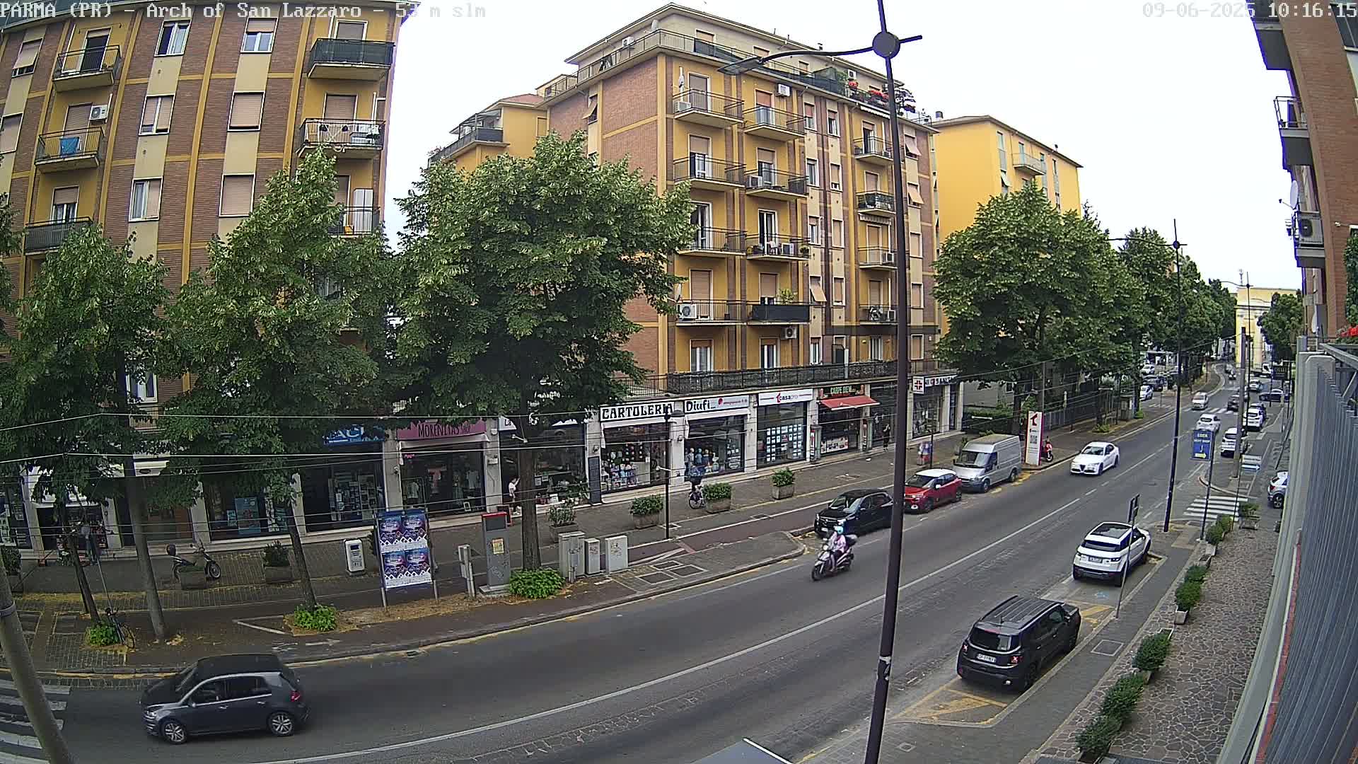 A street scene on an overcast day shows a multi-story building with shops on the ground floor, and cars parked and driving along the road in front of it.