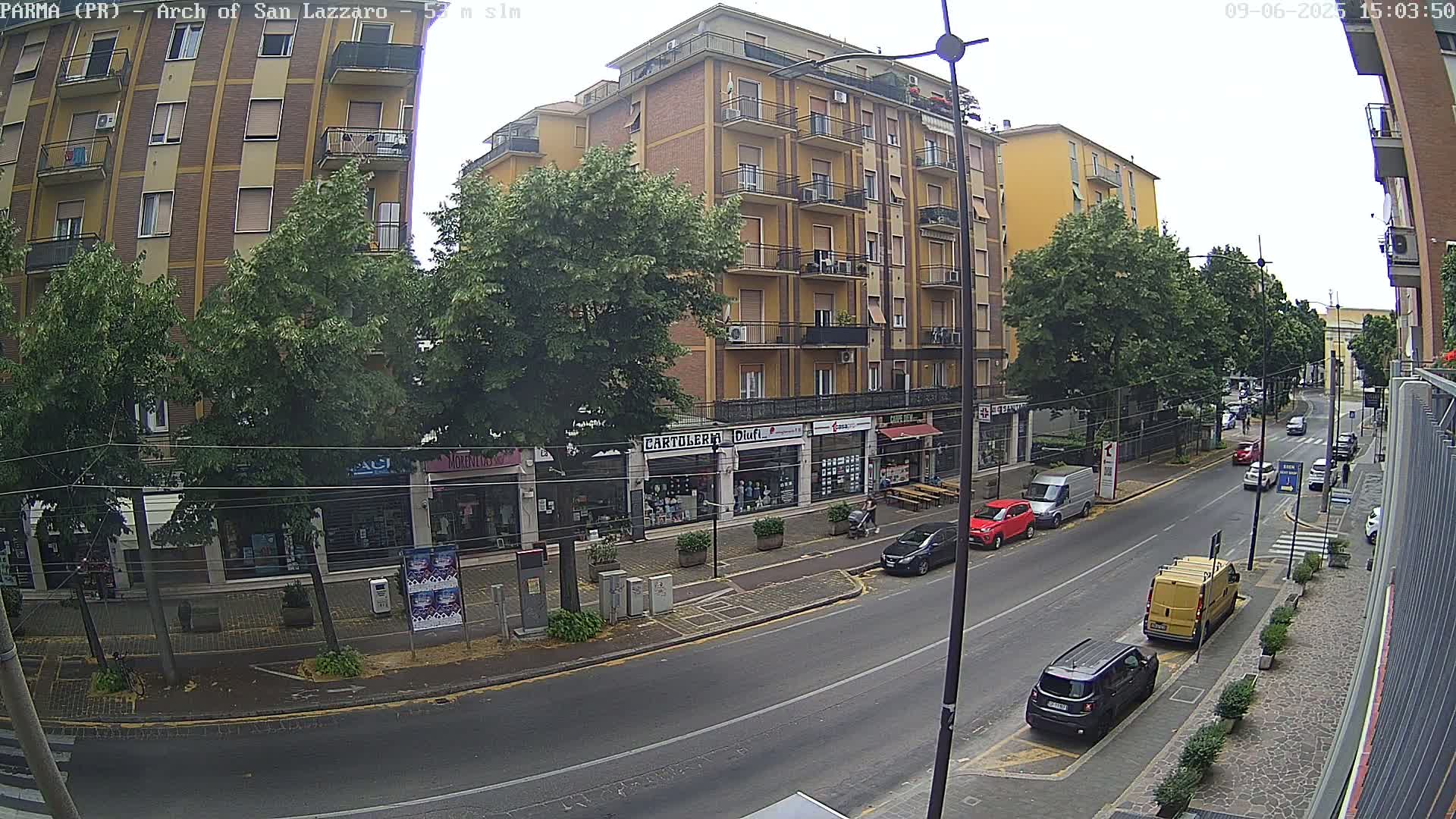 A street scene on an overcast day shows a multi-story building, shops, parked cars, and a few cars driving down the street.