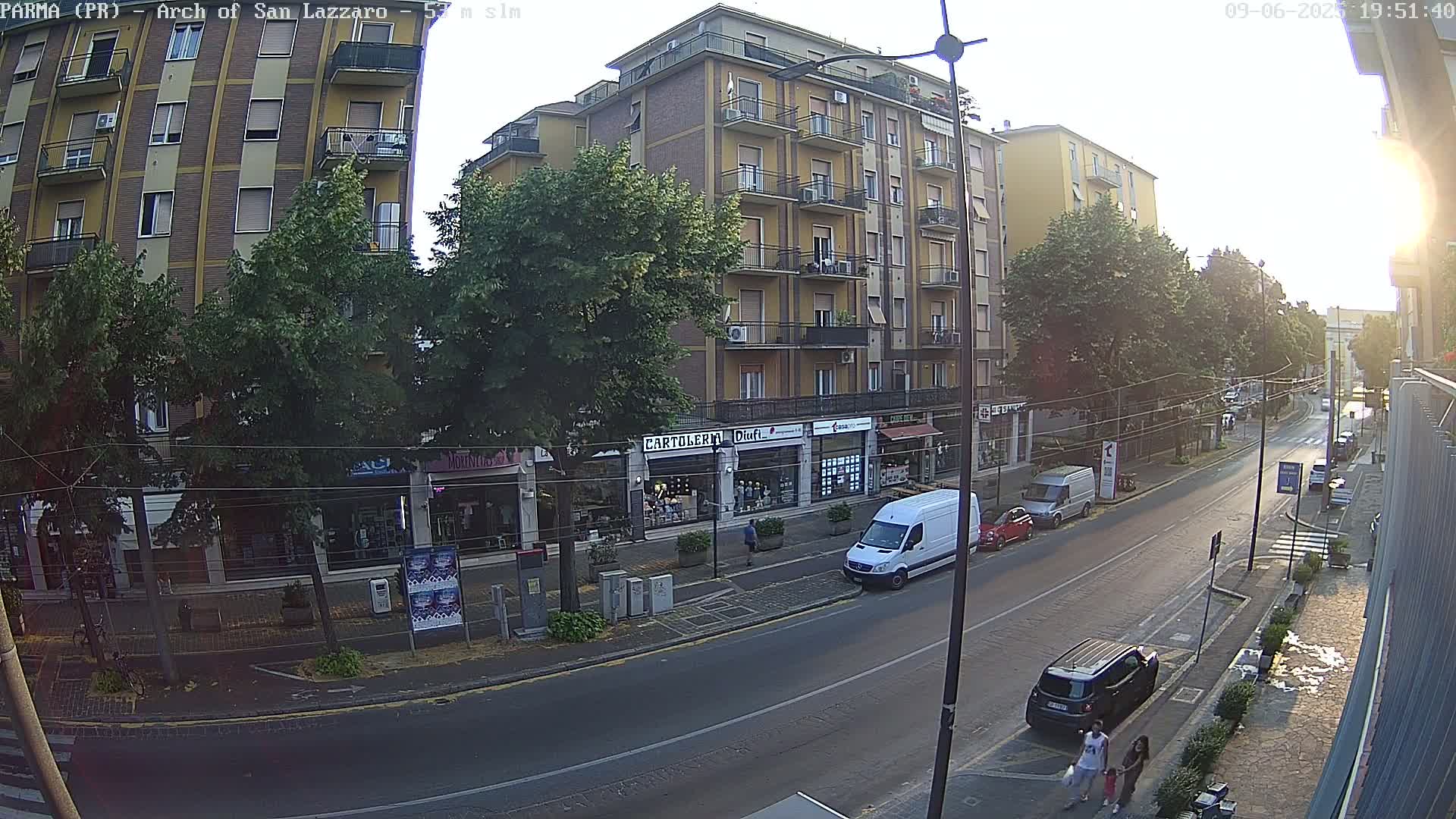 A street scene at sunset shows a multi-story building with shops on the ground floor, and cars parked along the road.