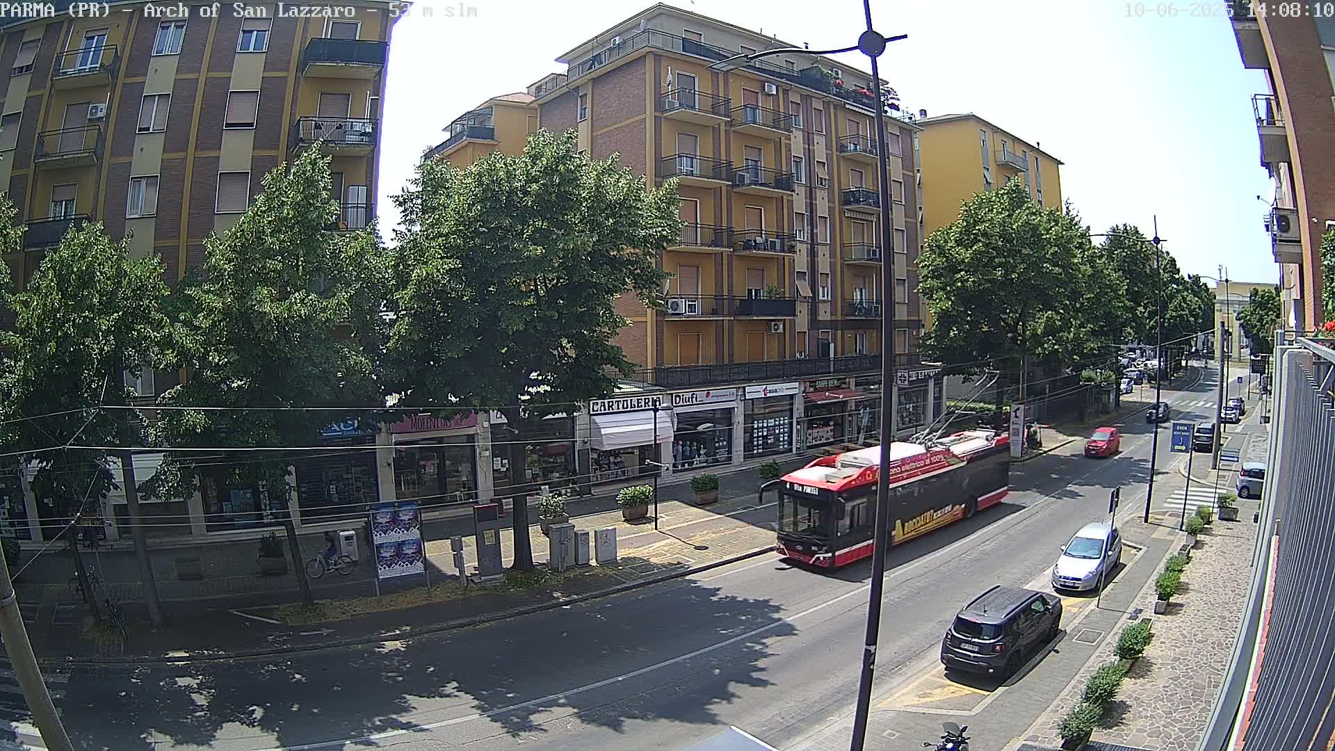 A sunny city street scene shows a red trolley bus driving past multi-story buildings and parked cars.