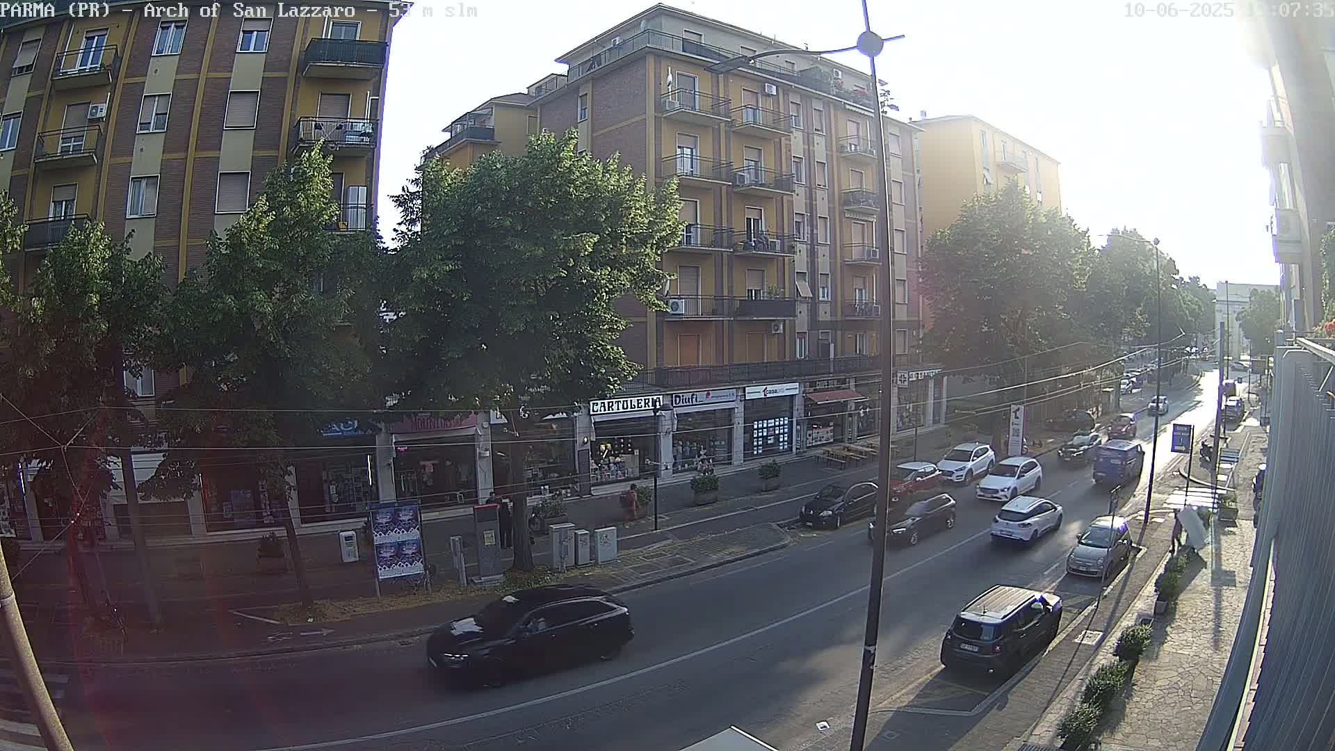 A sunny street scene shows a multi-story building with shops on the ground floor, several cars driving on the road in front of it, and some pedestrians on the sidewalk.