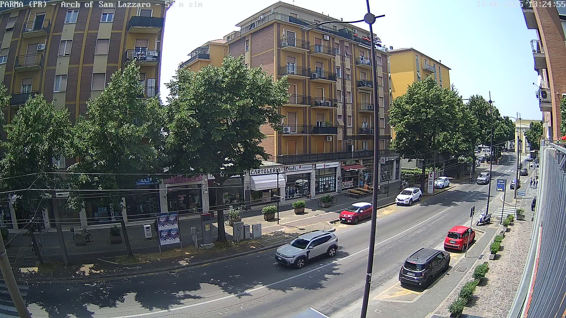 A sunny day reveals a street scene with several cars parked and driving along a road lined with shops and a multi-story building, all under a bright, clear sky.