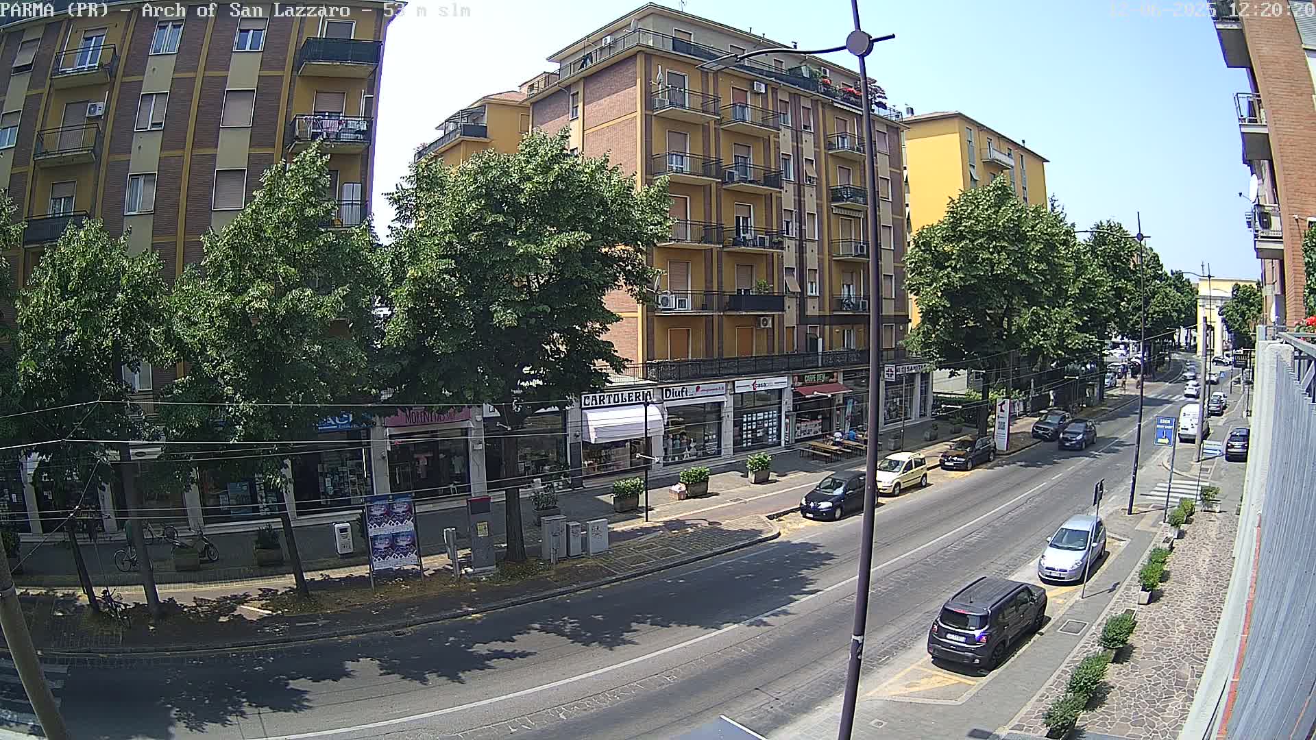 A sunny street scene shows a multi-story building with shops on the ground floor, lined with trees, and several cars parked and driving along a road.