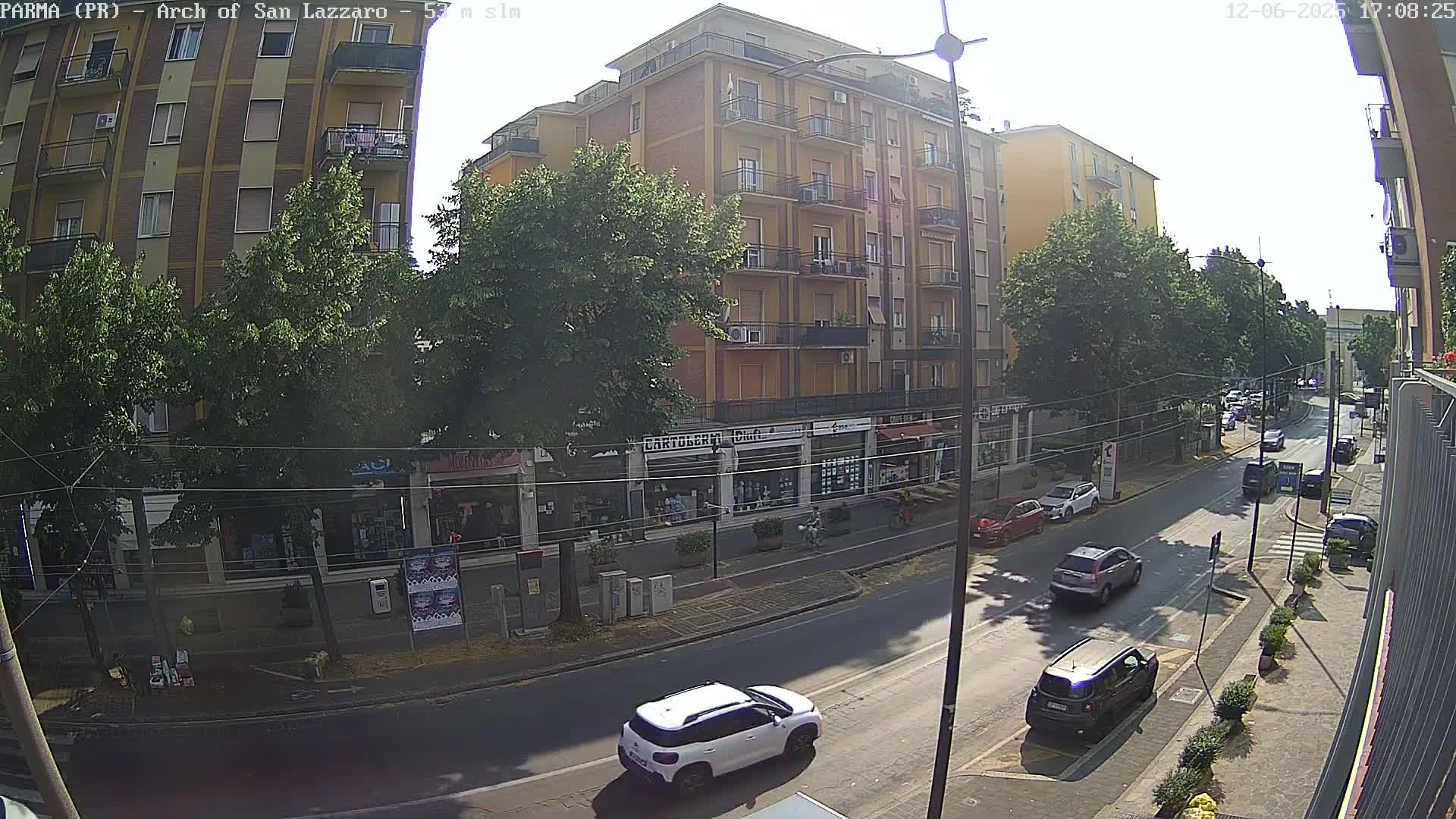 A sunny street scene shows a multi-story building with shops on the ground floor, cars parked and driving on the road in front, and trees lining the street.