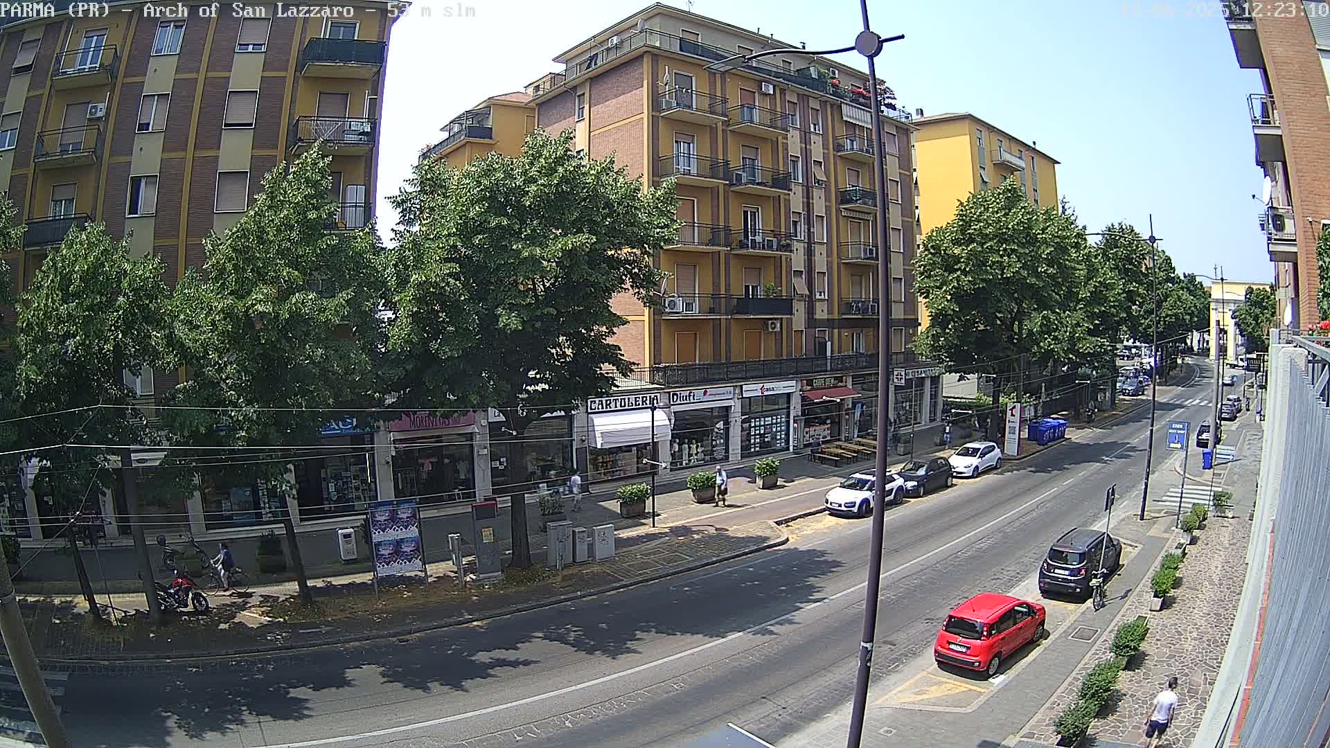 A sunny street scene shows a multi-story building with shops on the ground floor, several parked cars, and pedestrians walking along a roadway with parked cars.