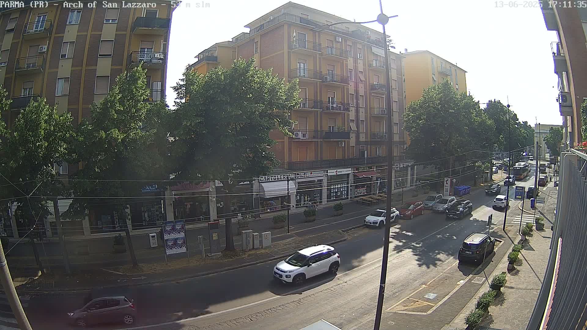 A sunny street scene shows a multi-story building with shops on the ground floor, lined with trees, with several cars parked and driving along the road.
