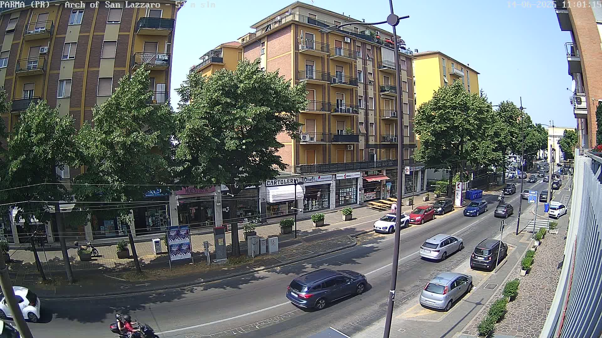 A sunny city street scene shows several cars parked and driving along a roadway lined with trees and shops, including a multi-story yellow building.