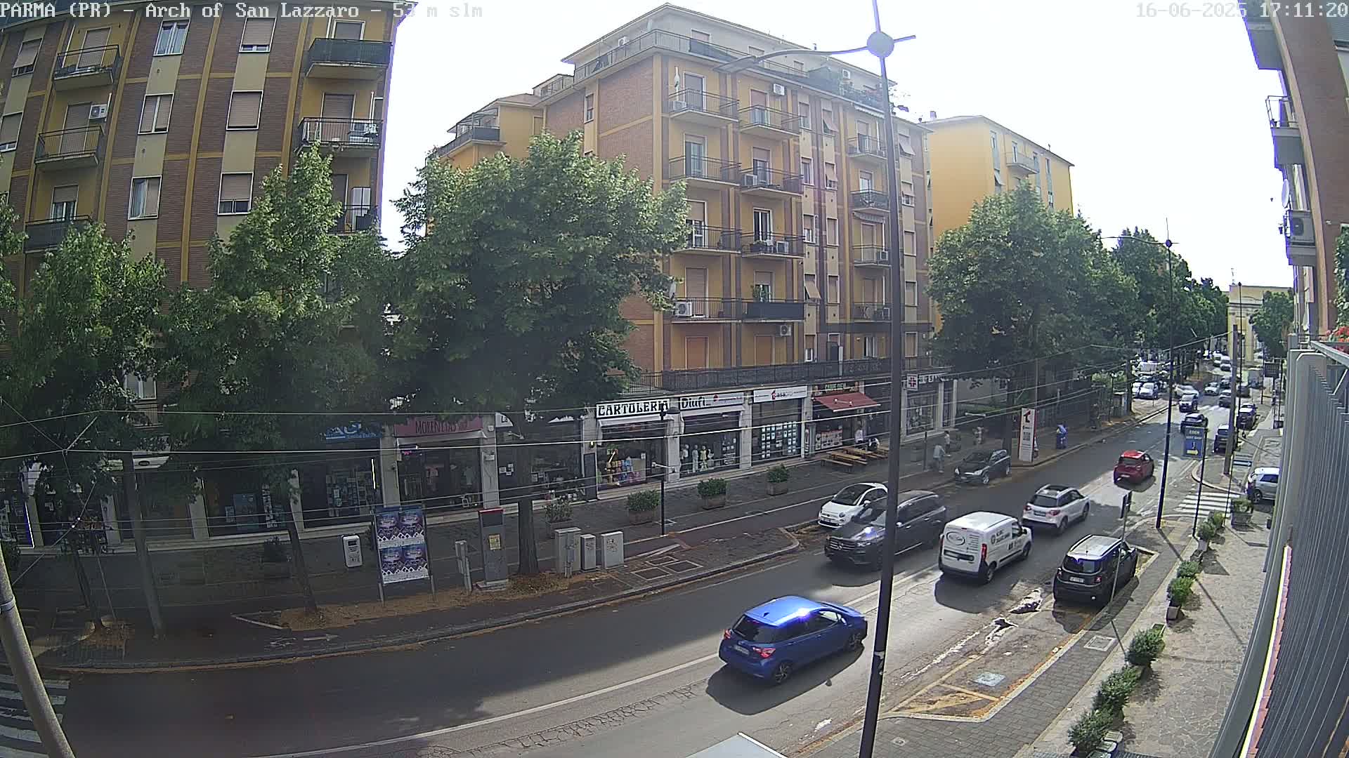 A sunny street scene shows a multi-story building with shops on the ground floor, lined with trees, and several cars driving down a road.