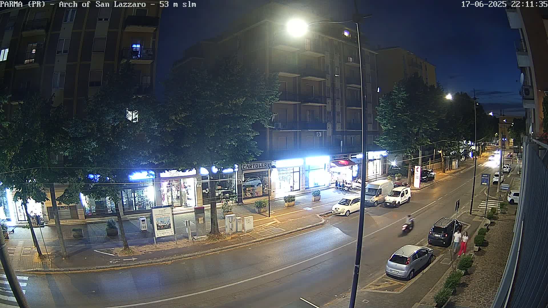 A nighttime street scene shows a road with parked cars and a few pedestrians, under streetlights, with buildings and trees lining both sides.
