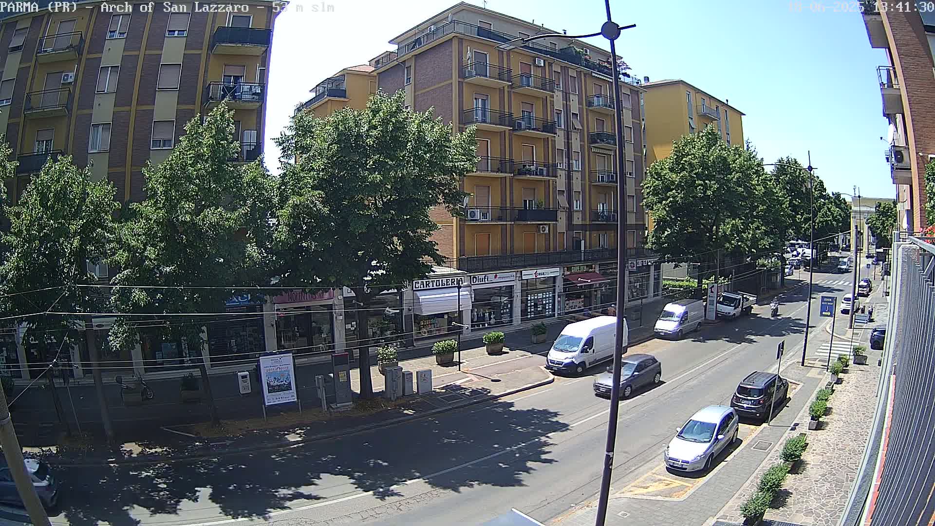 A sunny day reveals a city street lined with multi-story buildings and parked cars, with several vehicles in motion.