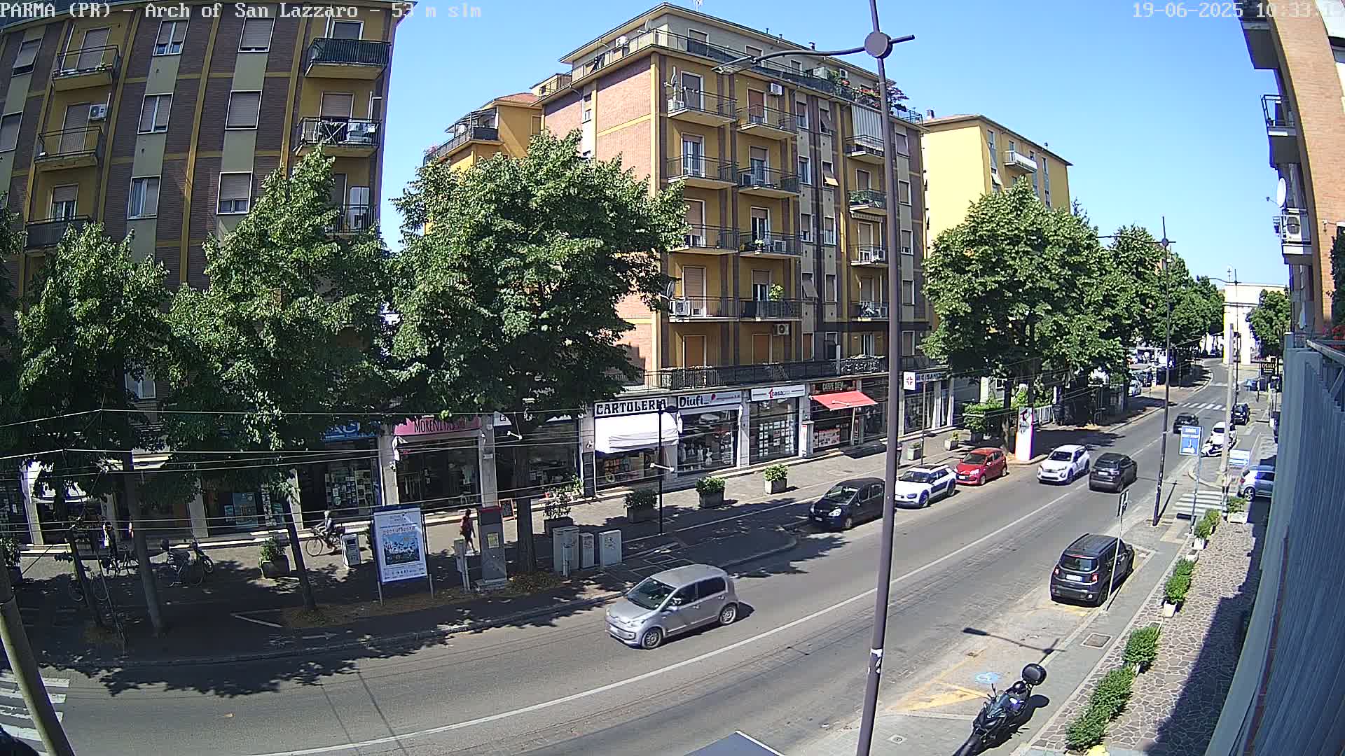 A sunny street scene shows a multi-story building with shops on the ground floor, lined with trees, and several cars parked and driving along the road.