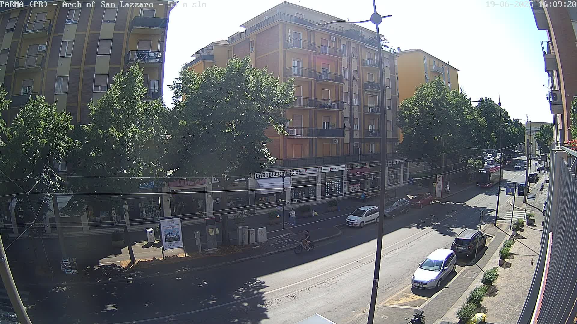 A sunny street scene shows a multi-story building, several parked cars, and a few pedestrians and cyclists on a city street lined with trees.