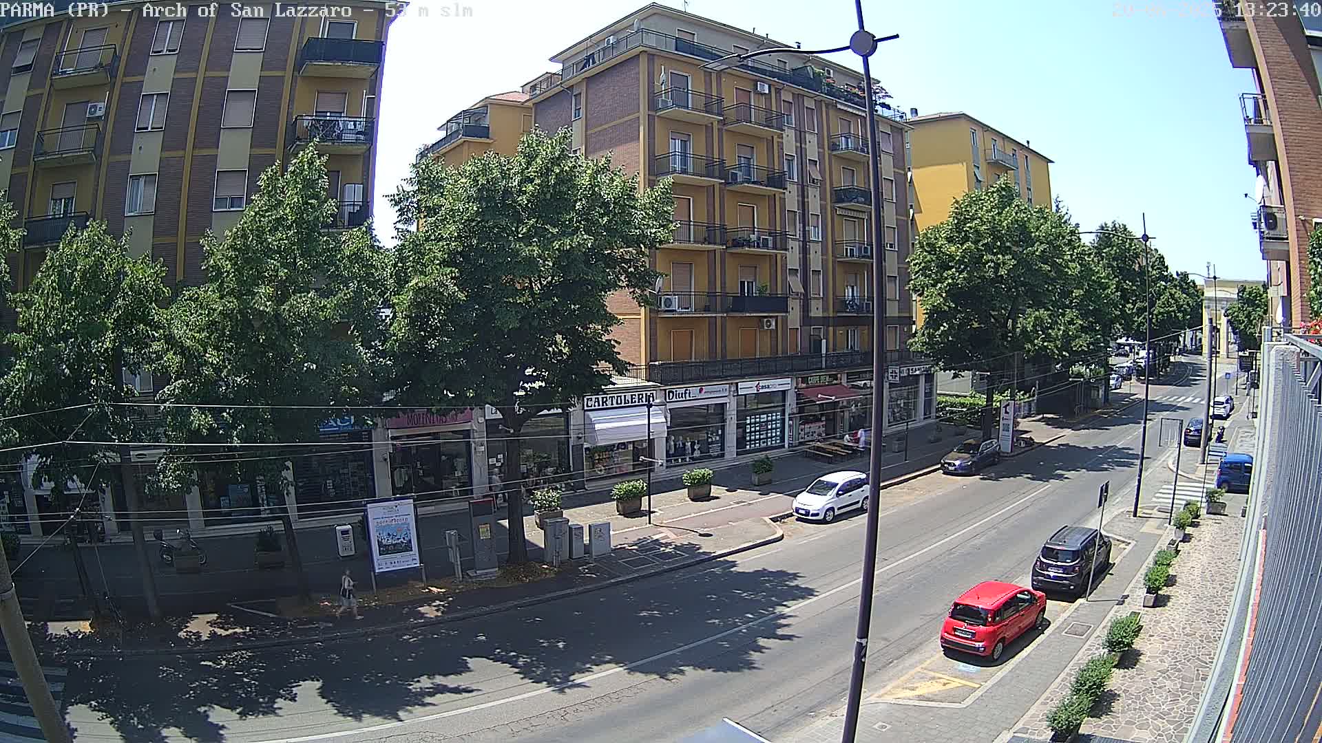 A sunny street scene shows a multi-story building with shops on the ground floor, lined with trees and parked cars, under a clear blue sky.