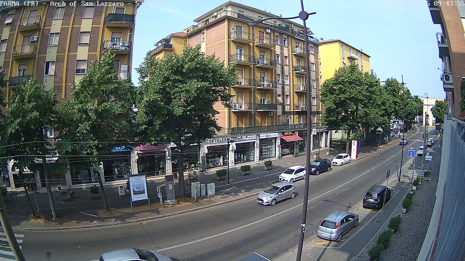 A sunny day reveals a street scene with several cars parked and driving past multi-story buildings and shops, lined with green trees.