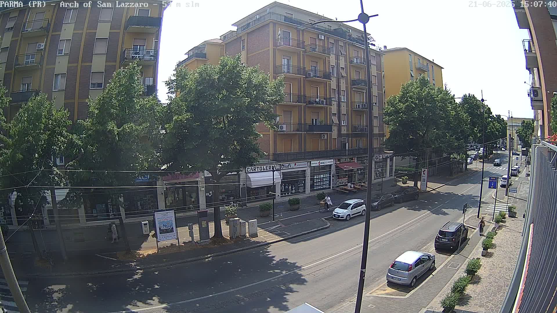 A sunny street scene shows a multi-story building with shops on the ground floor, parked cars, and pedestrians walking along a roadway lined with trees.