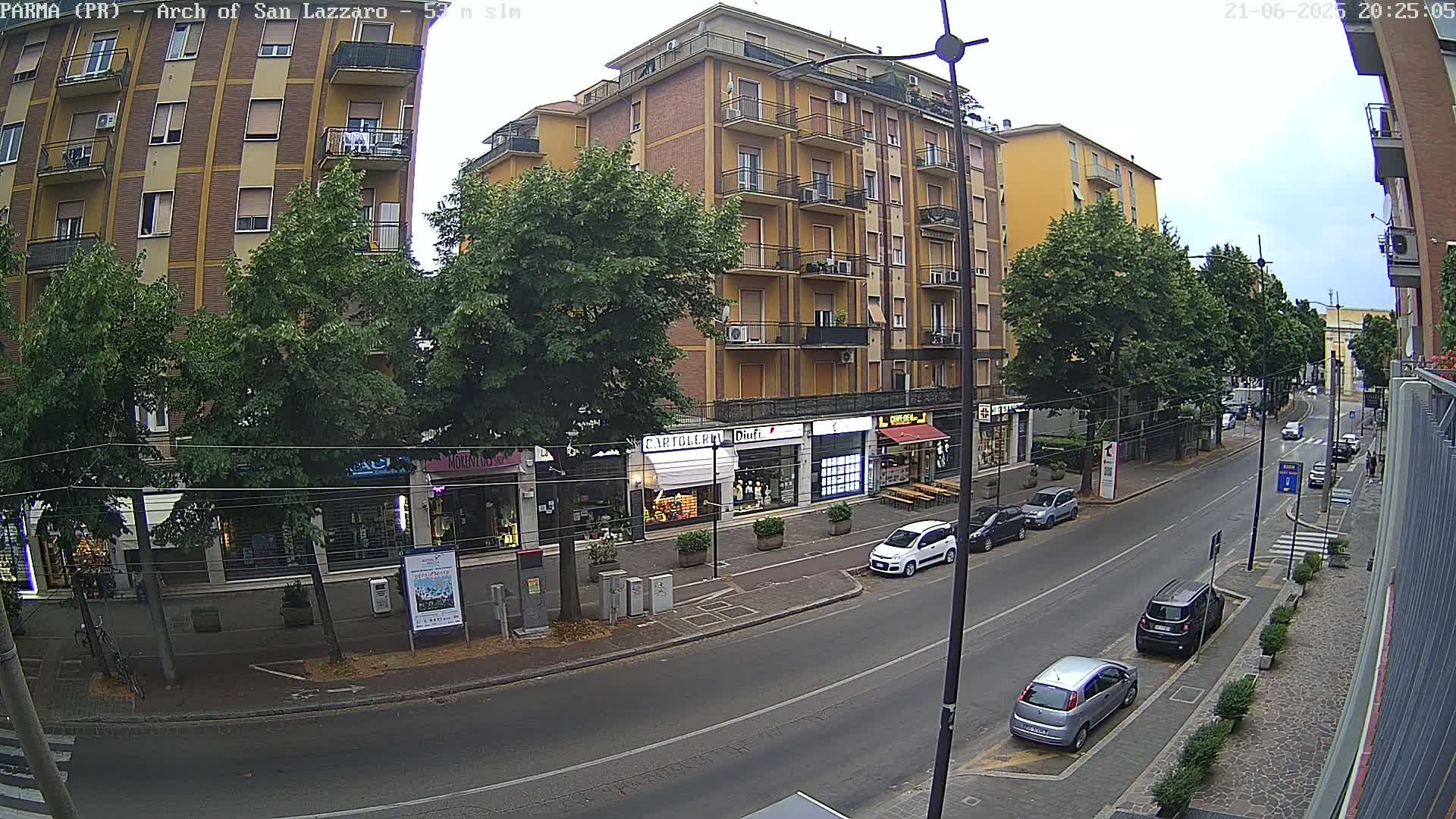 A street scene on an overcast day shows a multi-story building with shops on the ground floor, lined with trees, and several cars parked along the street and driving on the road.