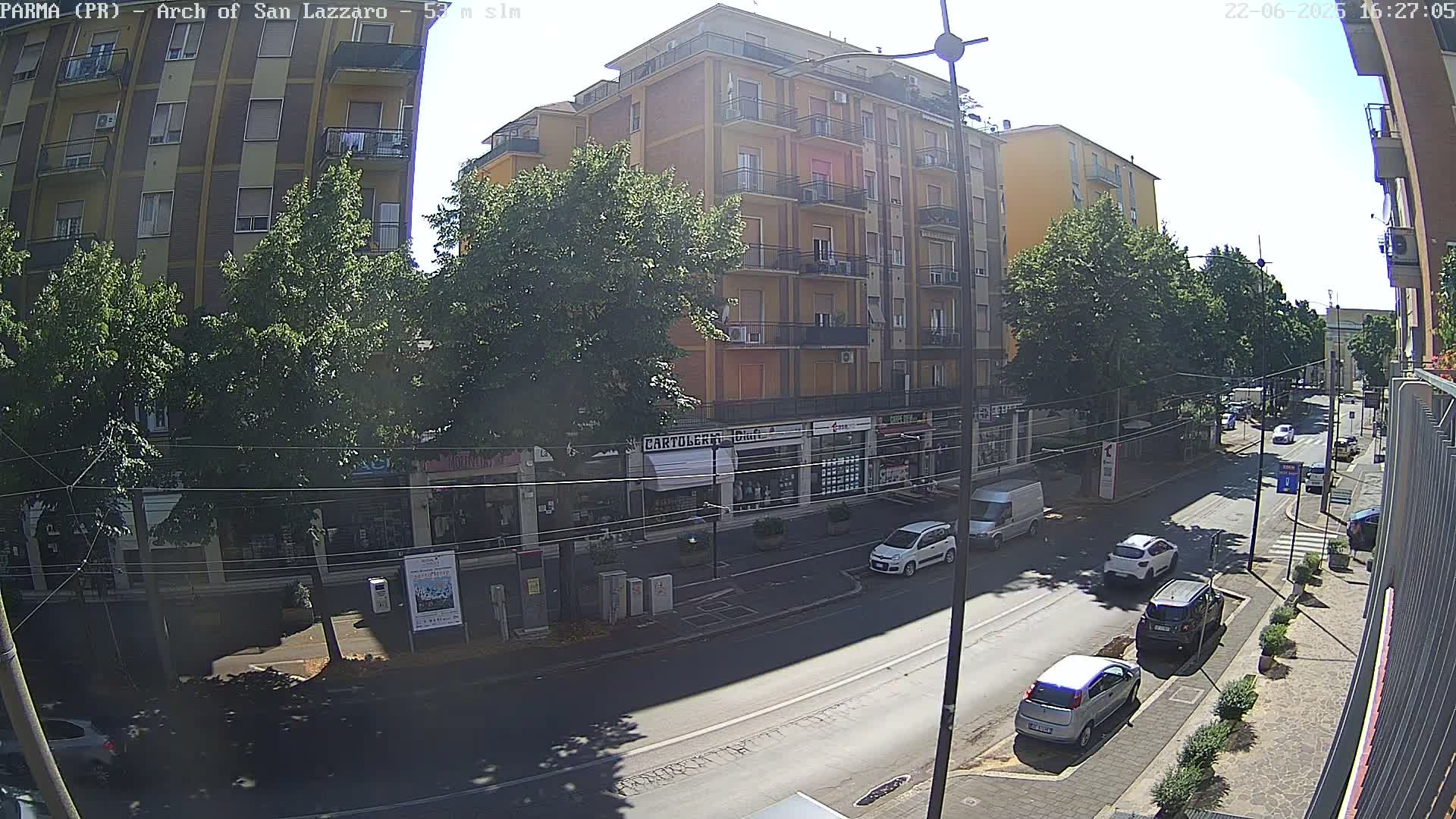 A sunny street scene shows a multi-story building with shops on the ground floor, several parked and moving cars, and lush green trees lining the street.