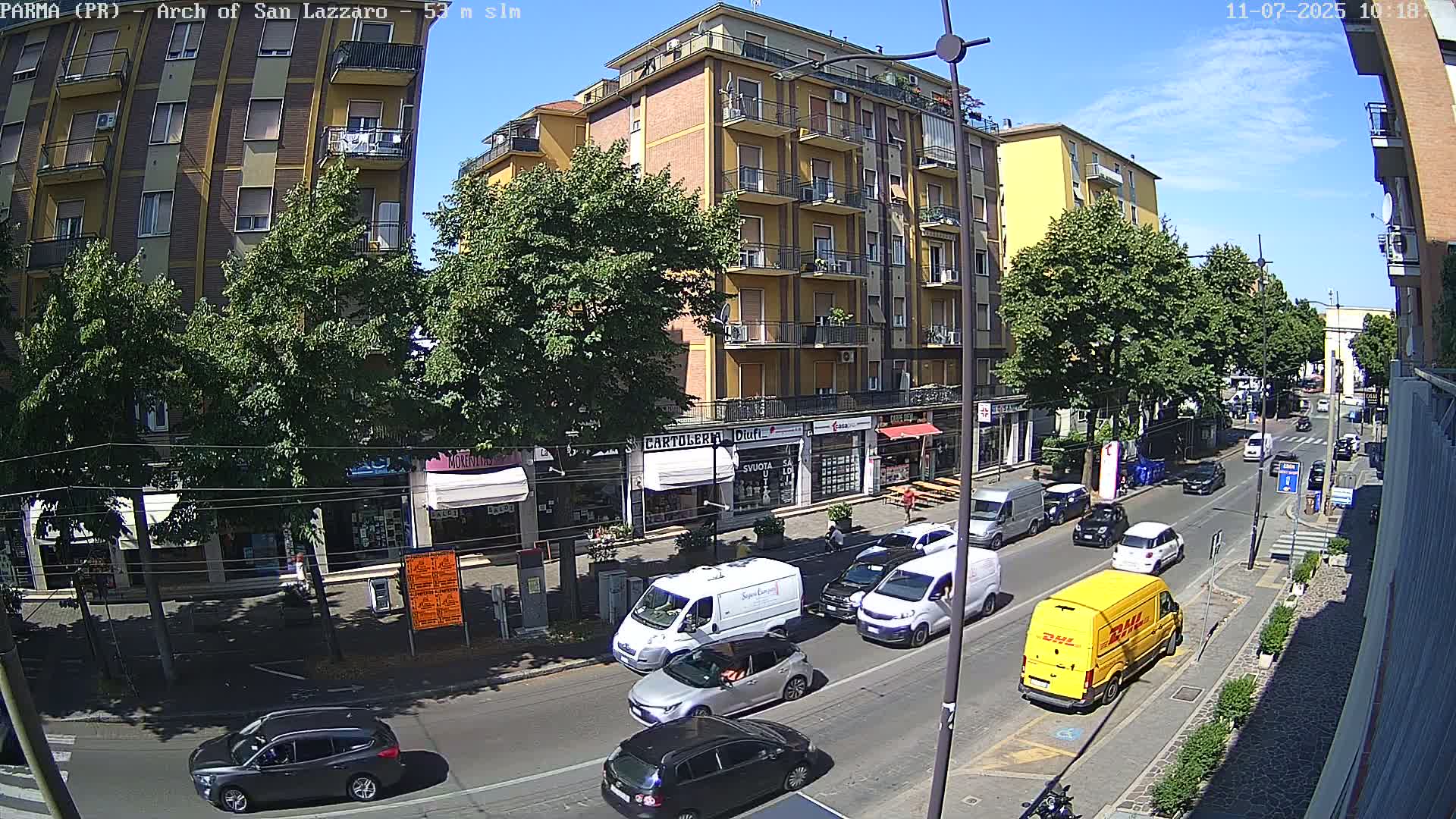 A sunny day on a city street with several cars and vans in motion, lined with buildings and trees.