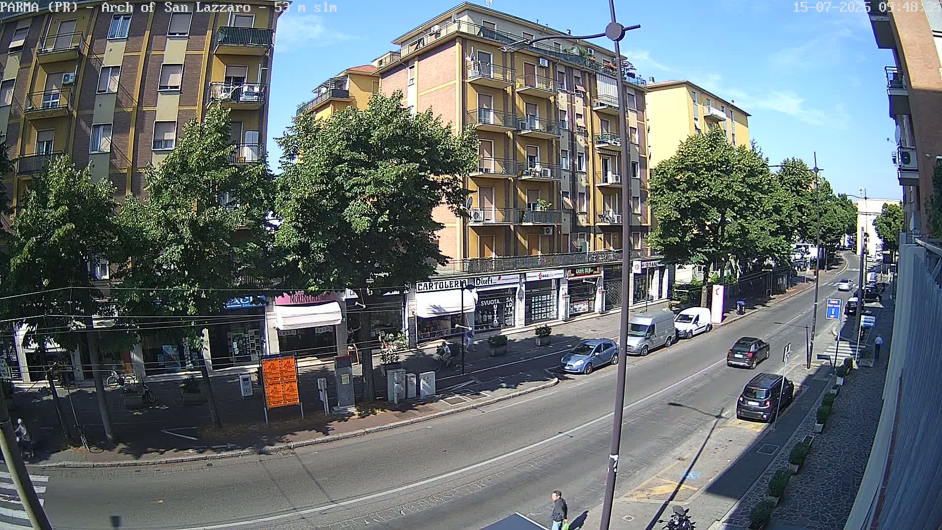 A sunny city street scene shows a multi-story building, cars parked along the side, and several cars driving down the street, with some pedestrians on the sidewalk.