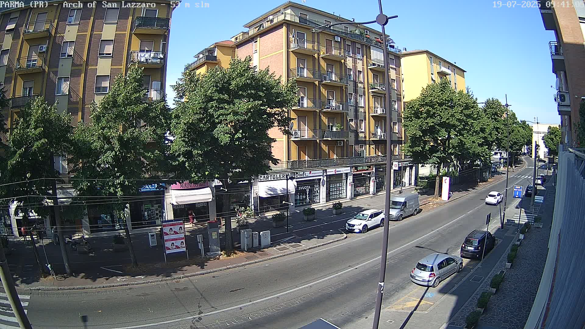 A sunny city street scene shows a multi-story yellow building and several parked cars, with a few cars driving on the road lined with trees and shops.