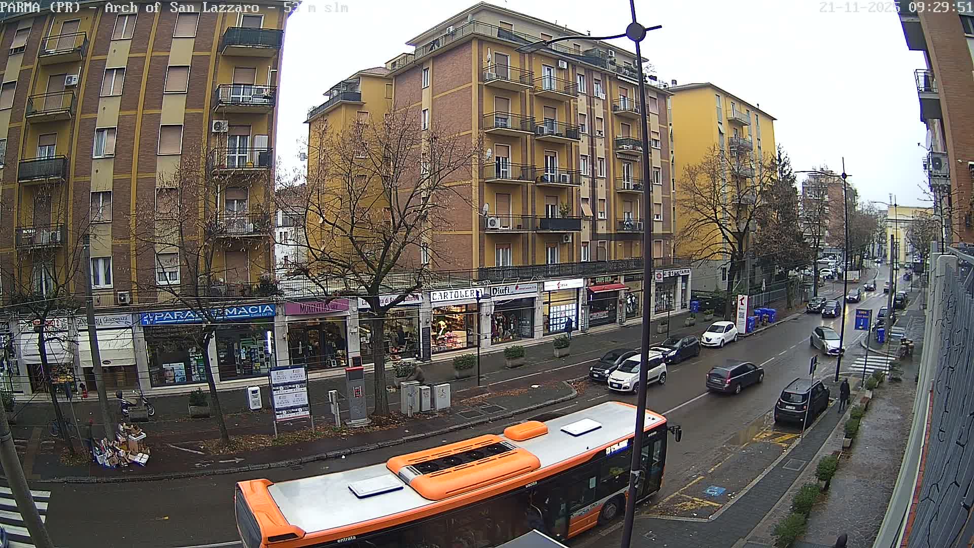 An overcast and wet urban street scene shows multi-story buildings, bare trees, and numerous vehicles including a prominent orange and grey bus in the foreground.