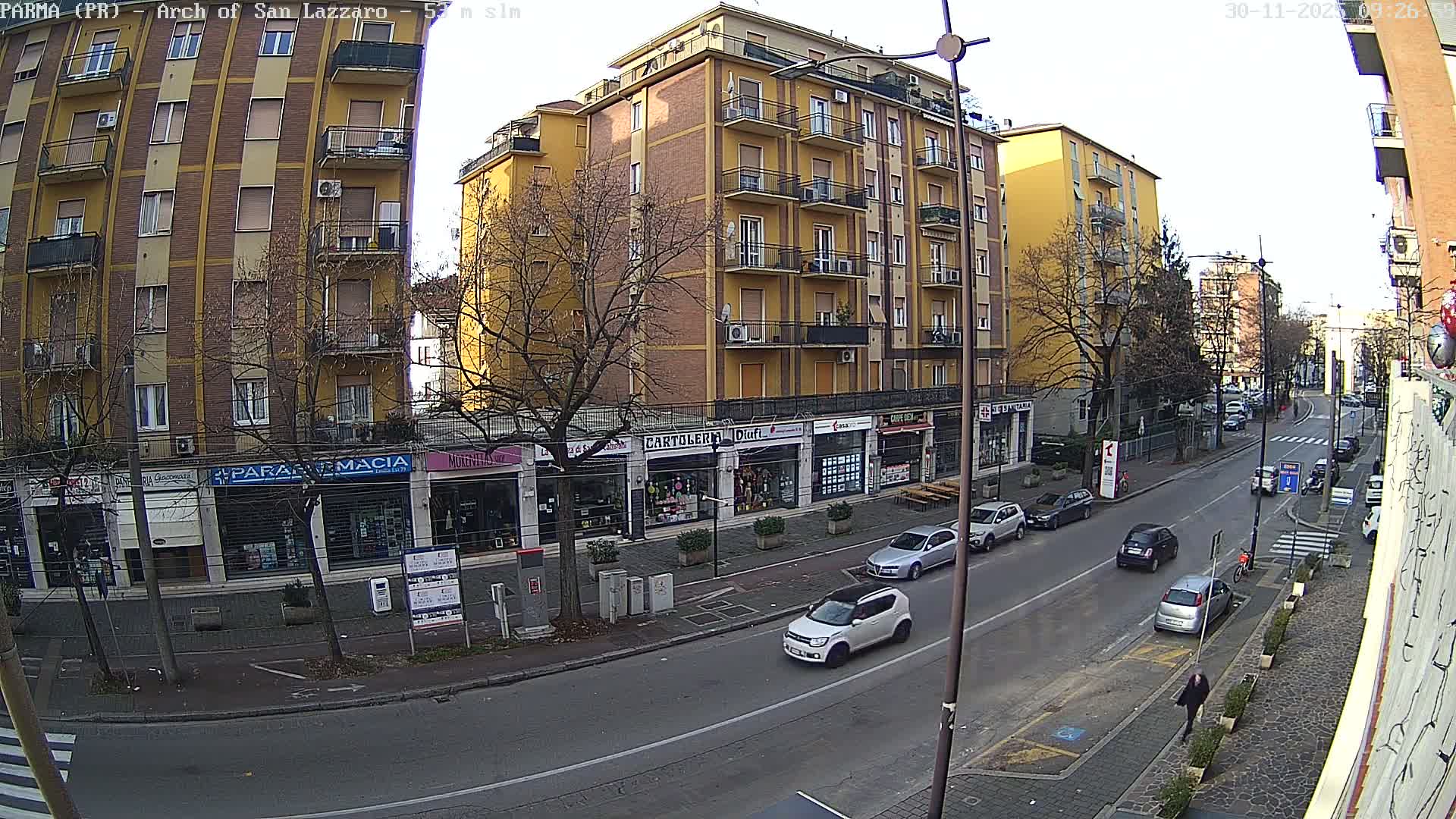 An overcast urban scene captures multi-story buildings with ground-floor shops, bare trees, and cars moving on a wet street while a few pedestrians walk along the sidewalks.