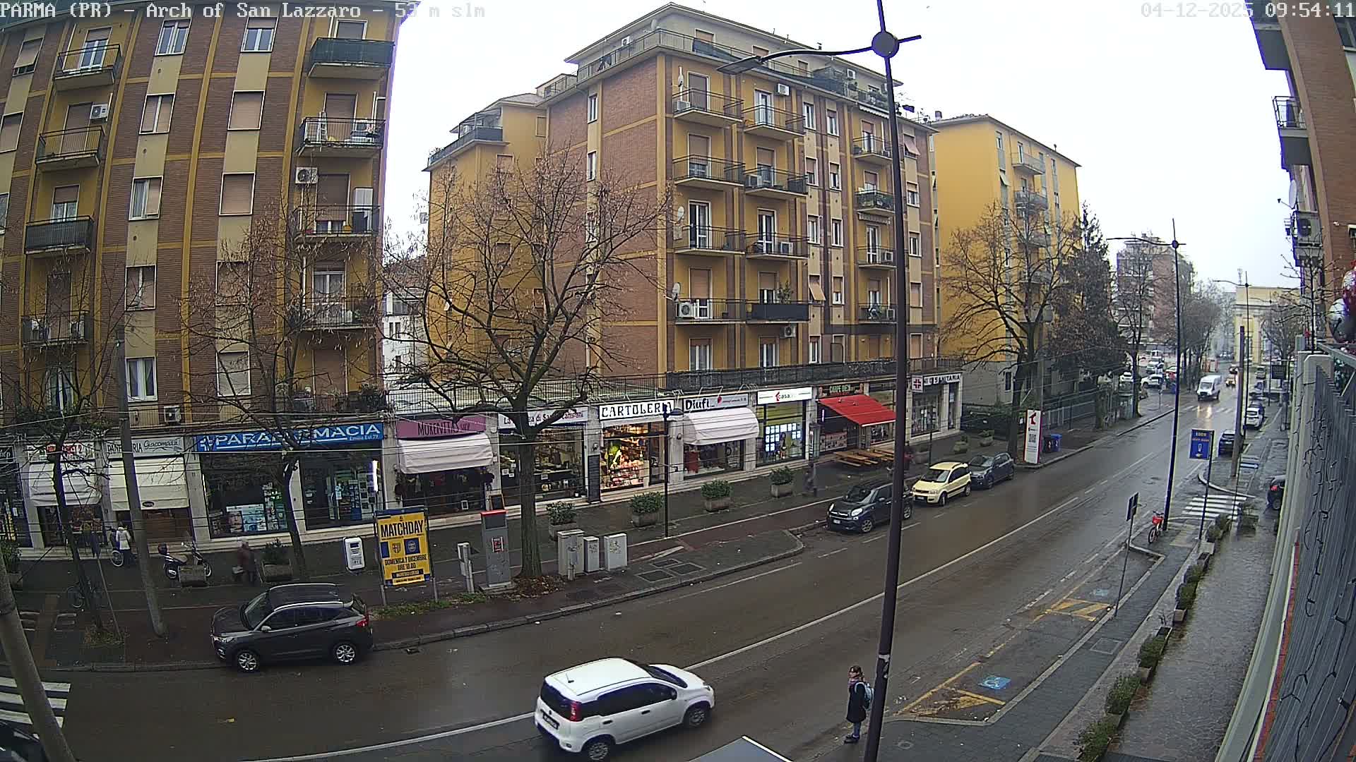 A wet city street, lined with multi-story buildings housing ground-floor shops and apartment balconies, features moving and parked cars and bare trees under a dull, overcast sky.