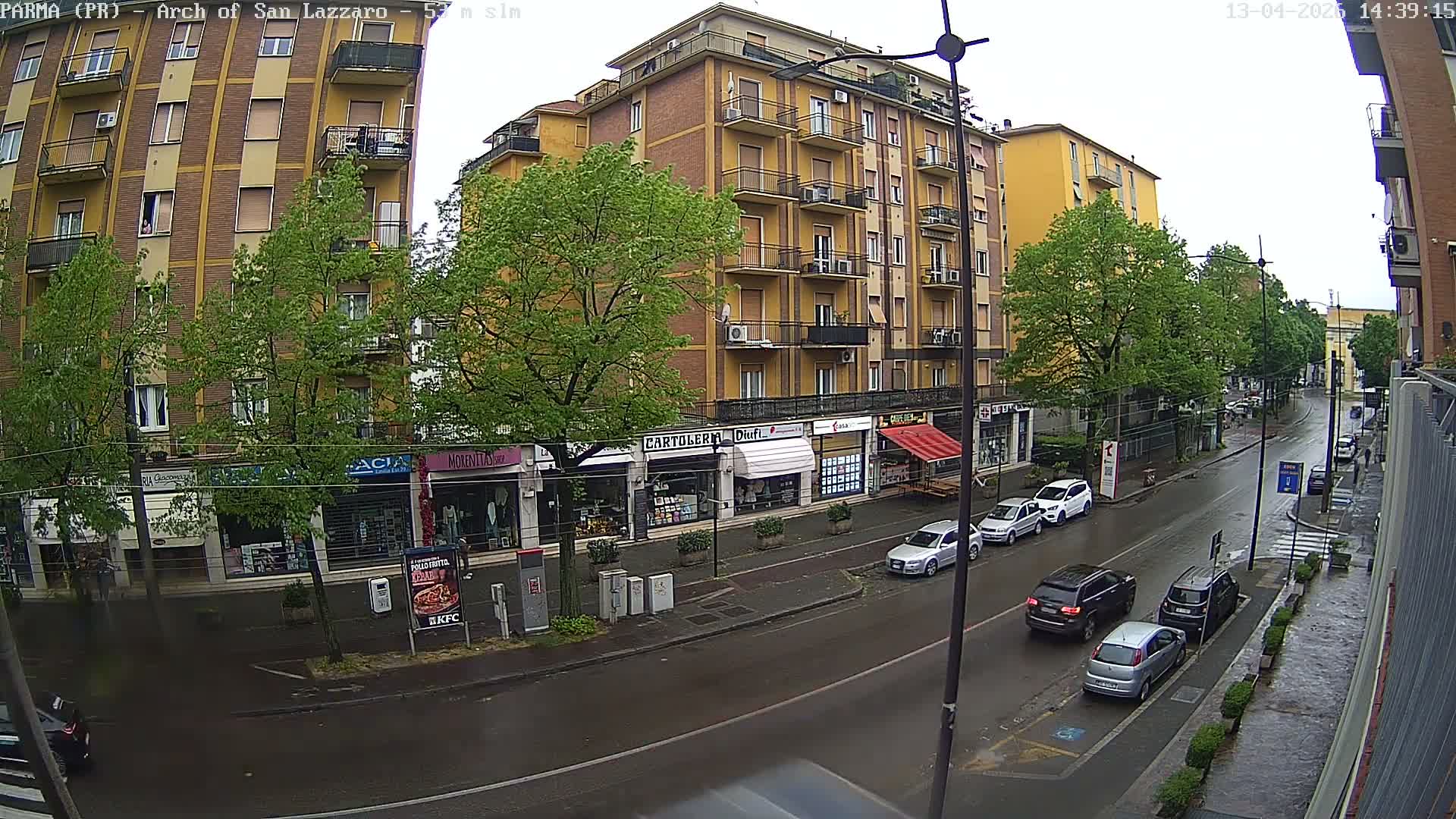 A sunny outdoor scene depicts a busy European city street lined with multi-story buildings housing ground-floor shops, bare trees, several parked and moving cars, and a pedestrian on the sidewalk.