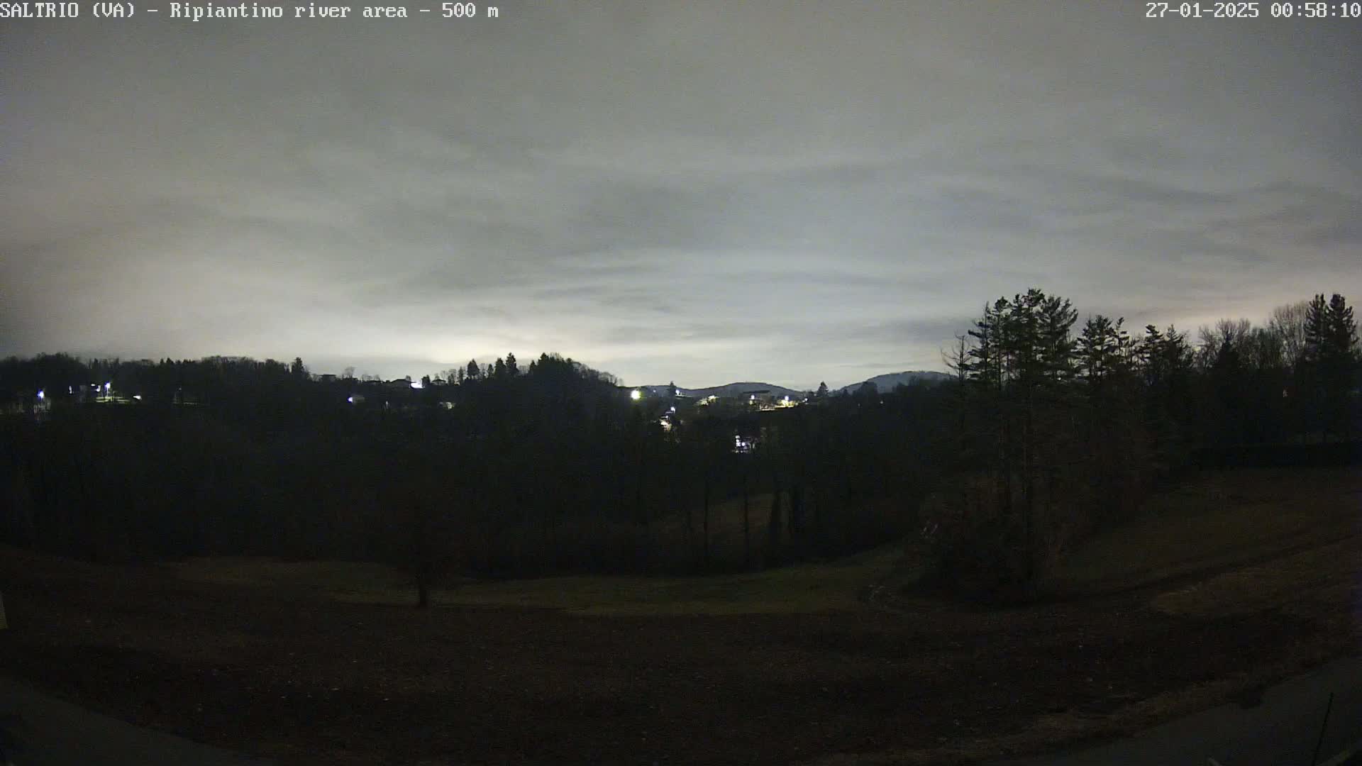 A nighttime view of a dark, hilly landscape with a few sparsely lit buildings in the distance under a cloudy sky.