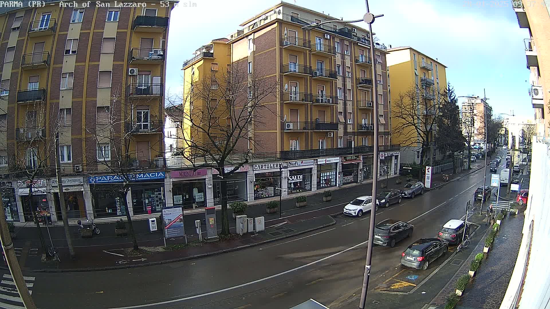 A wet city street scene shows several cars parked and driving past multi-story buildings with shops on the ground floor, under a mostly clear sky.